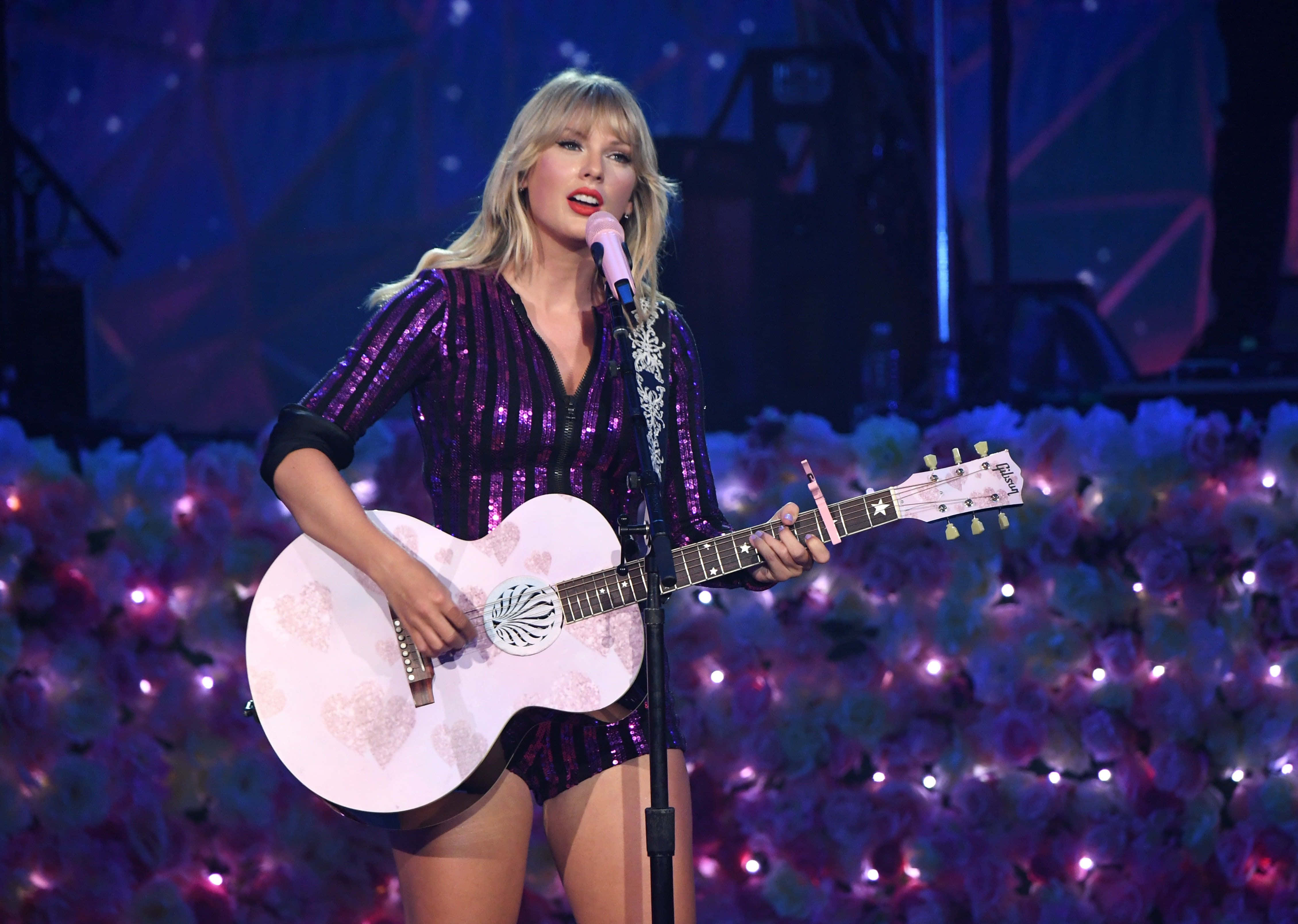 桌布 壁紙 — Taylor Swift 在紫色舞台燈光下手持白色花卉木吉他在舞台上表演。