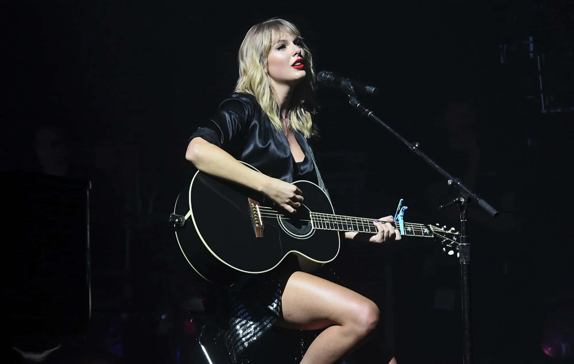 桌布 壁紙 — Taylor Swift 在舞台上手持黑色木吉他，在戲劇性的舞台燈光下表演。