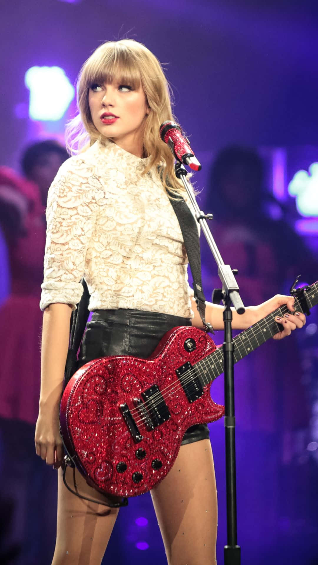 Taylor Swift iPhone 桌布 壁紙 — Taylor Swift 在舞台上表演，手持閃閃發光的紅色電吉他，在充滿活力的紫色舞台燈光下。
