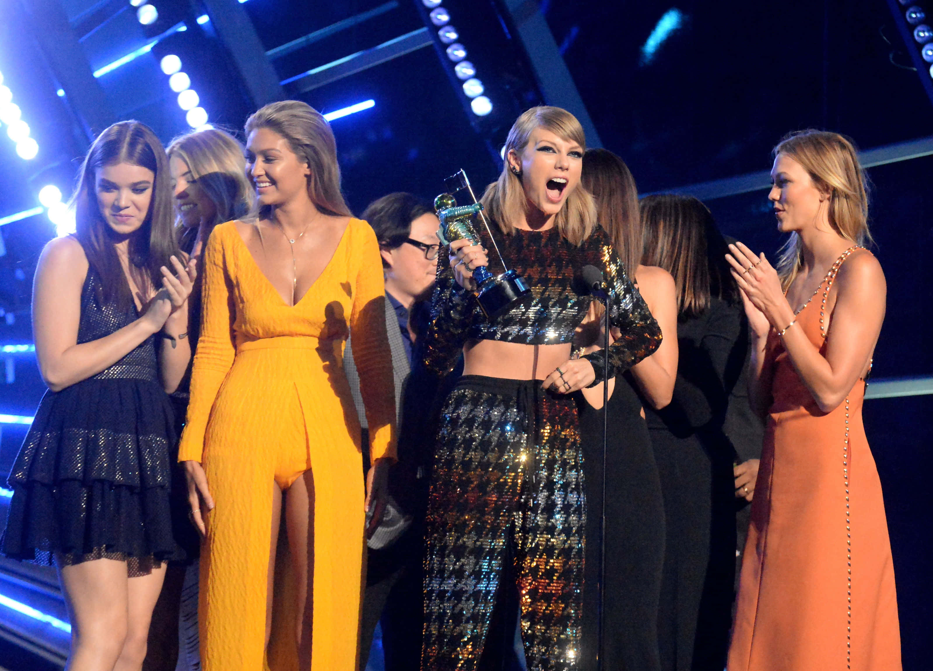 squad 桌布 壁紙 — Taylor Swift 在舞台上穿著閃亮服裝，手持獎項慶祝，朋友們在明亮的藍色舞台燈光下鼓掌。