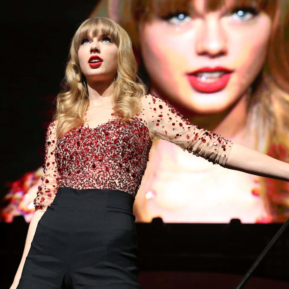 泰勒絲 桌布 壁紙 — Taylor Swift 在舞台上表演，身穿閃亮紅色緊身胸衣和黑色褲子，身後有巨大螢幕。