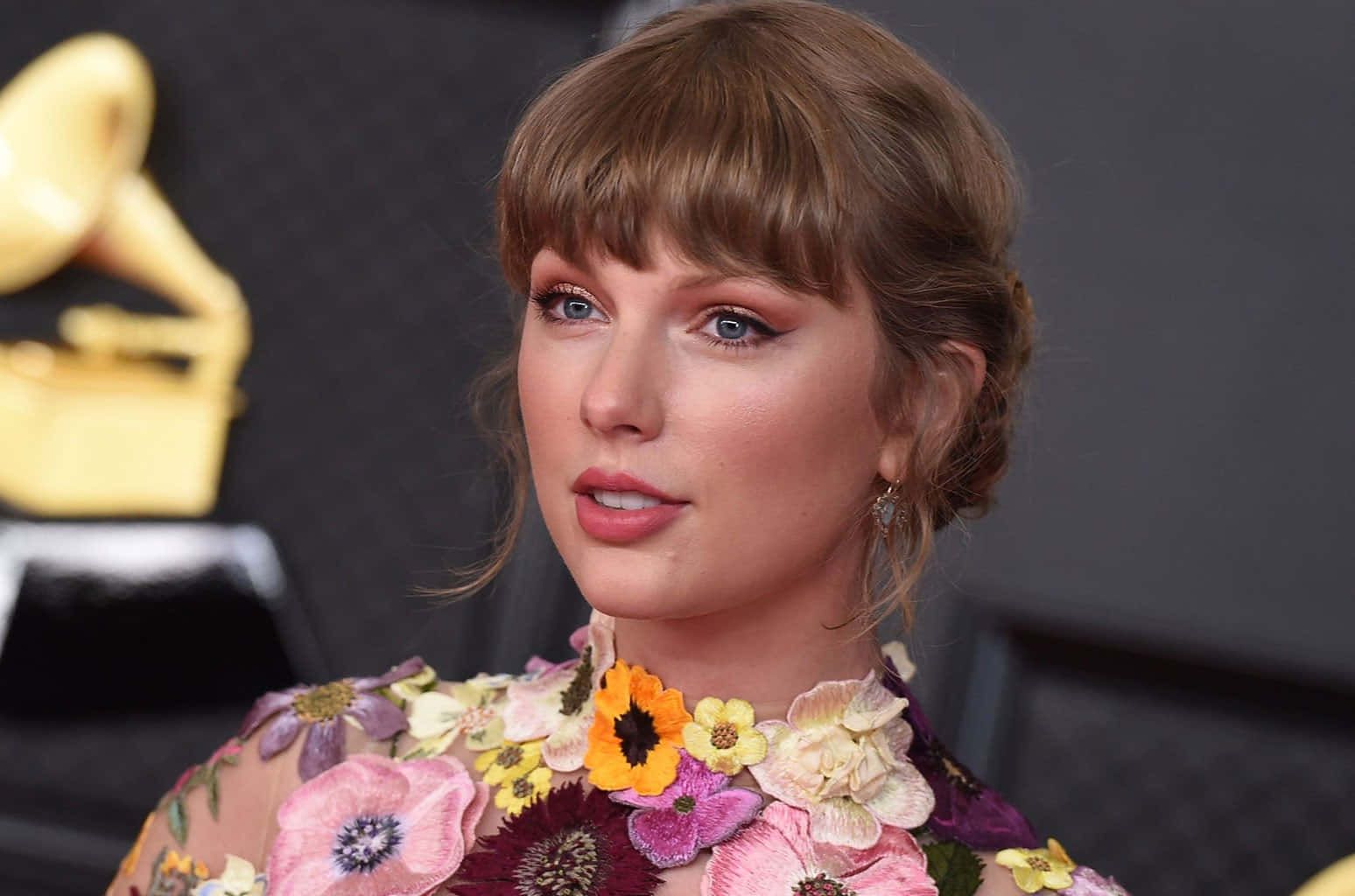 泰勒絲 桌布 壁紙 — Taylor Swift 溫柔地凝視，身穿花卉長裙，身後有模糊的葛萊美獎座。