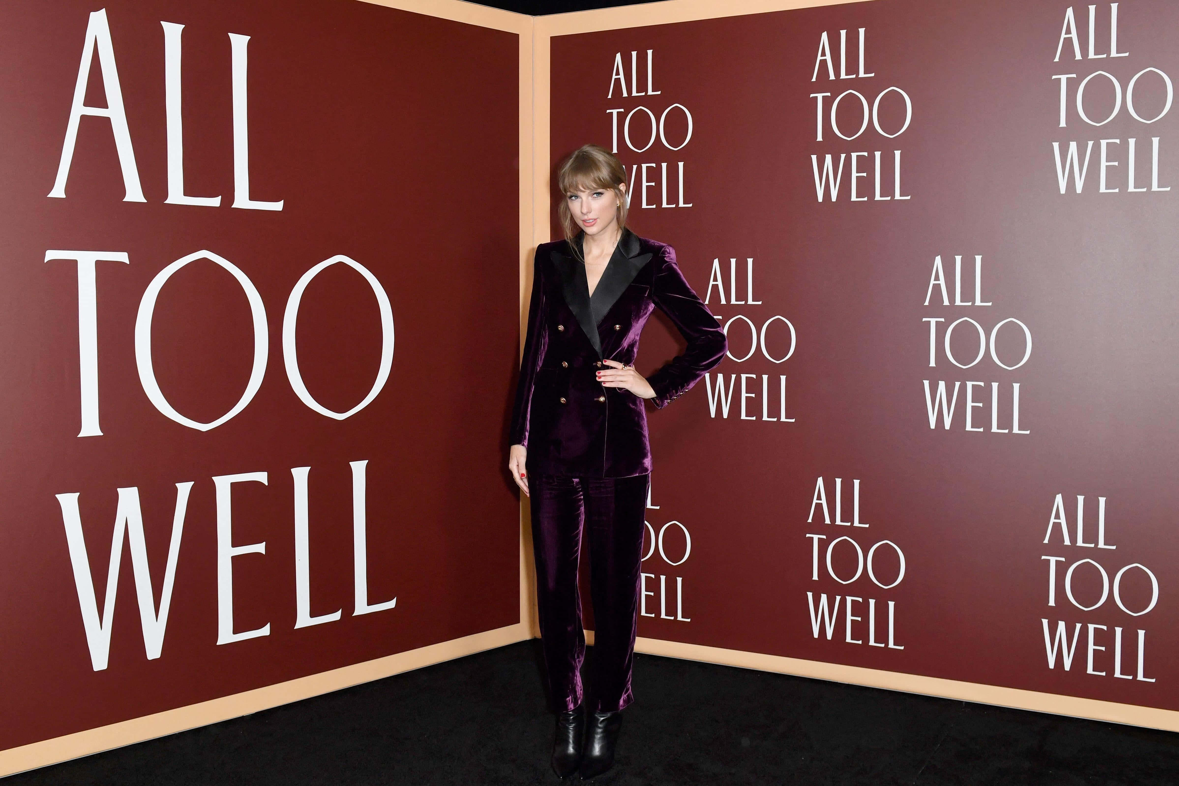 泰勒絲 桌布 壁紙 — Taylor Swift 自信地擺姿勢，身穿紫色天鵝絨西裝，背景是重複顯示「ALL TOO WELL」字樣的栗色牆壁。