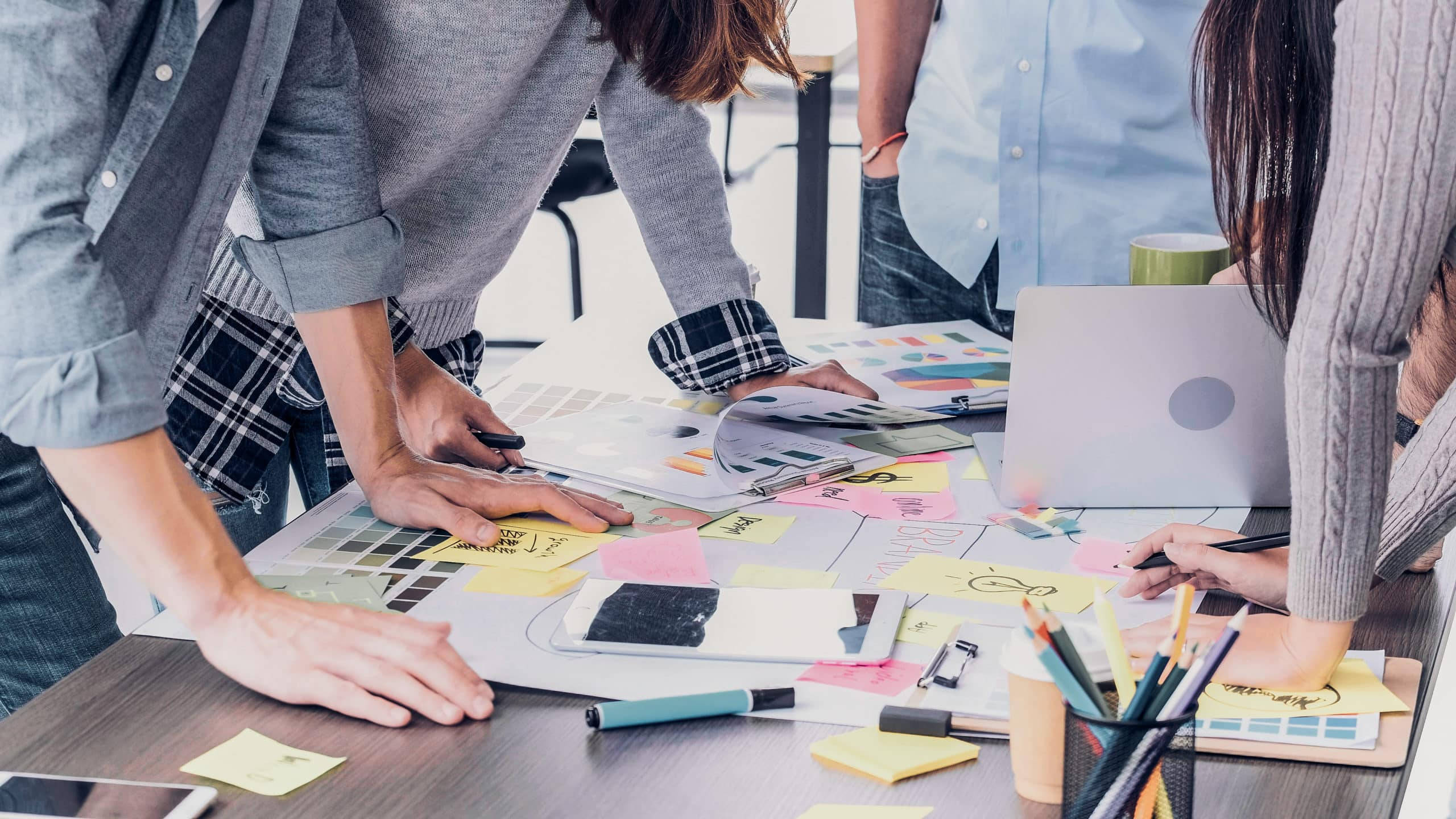 學校 桌布 壁紙 — Team members collaborate around a cluttered desk covered in colorful sticky notes design mockups and digital tools.