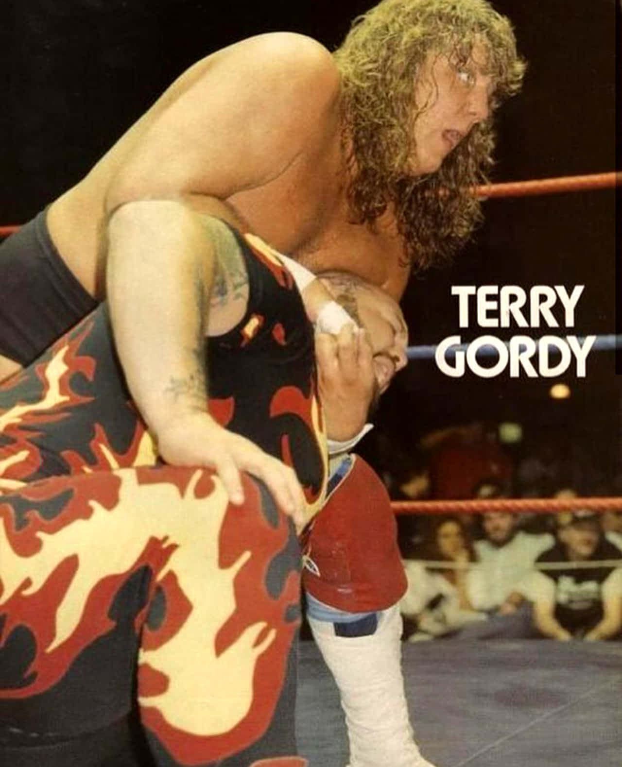 WWE 桌布 壁紙 — Terry Gordy 在明亮的場館燈光下，穿著火焰圖案緊身衣，在激烈的摔角比賽中舉起對手。