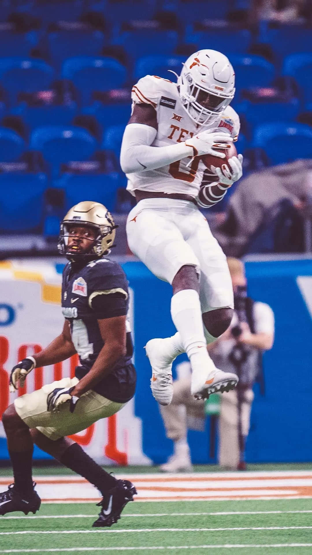  壁紙 — 一位德州足球員在半空中躍起緊抓著球，而一名防守球員在他下方撲向充滿活力的綠色球場。