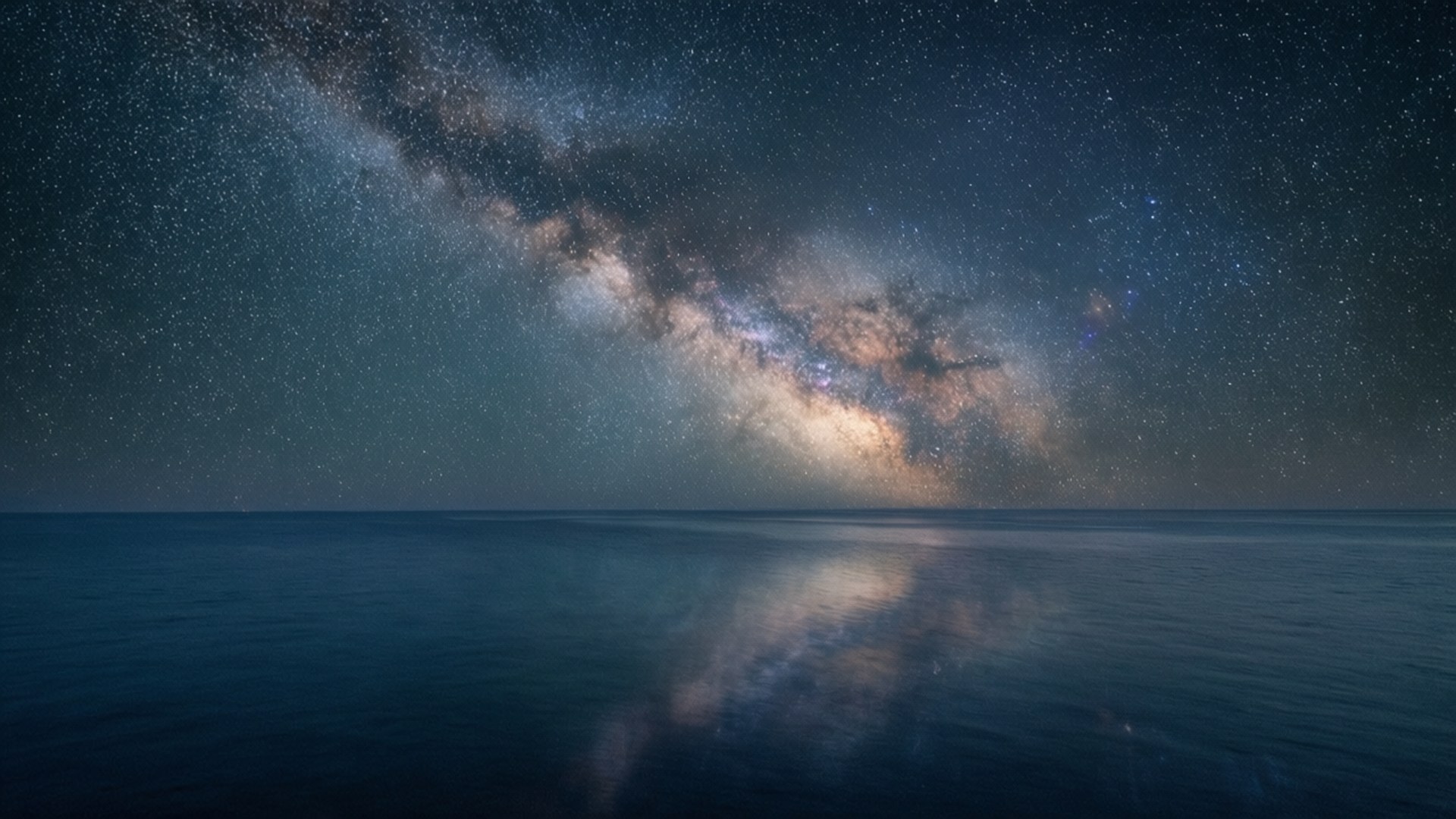 Milky Way Over Ocean 壁紙 — 銀河系在繁星點點的夜空中燦爛地彎曲，完美地倒映在平靜的海面上。