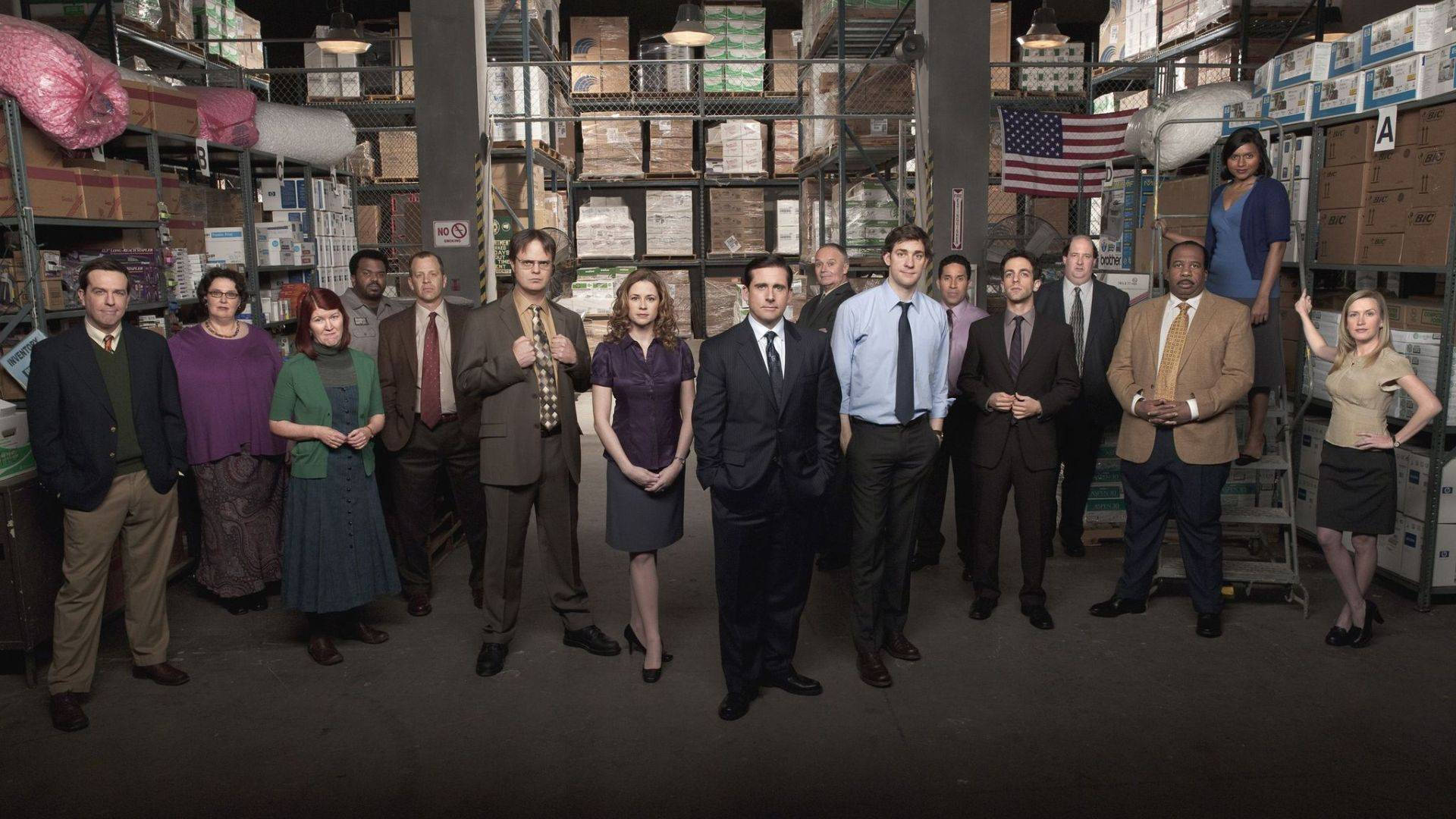 辦公室 桌布 壁紙 — 《The Office》演員在一個雜亂的倉庫中擺姿勢，倉庫裡擺滿了貨架上的盒子和一面美國國旗。