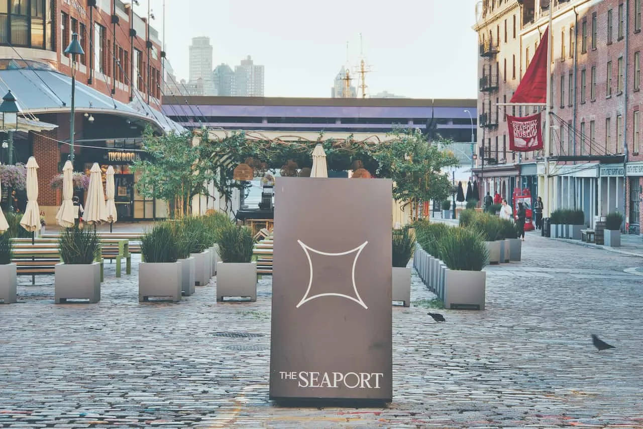 New York 桌布 壁紙 — The Seaport 的鵝卵石廣場設有現代花盆、關閉的雨傘和一個背景為城市天際線的標誌。