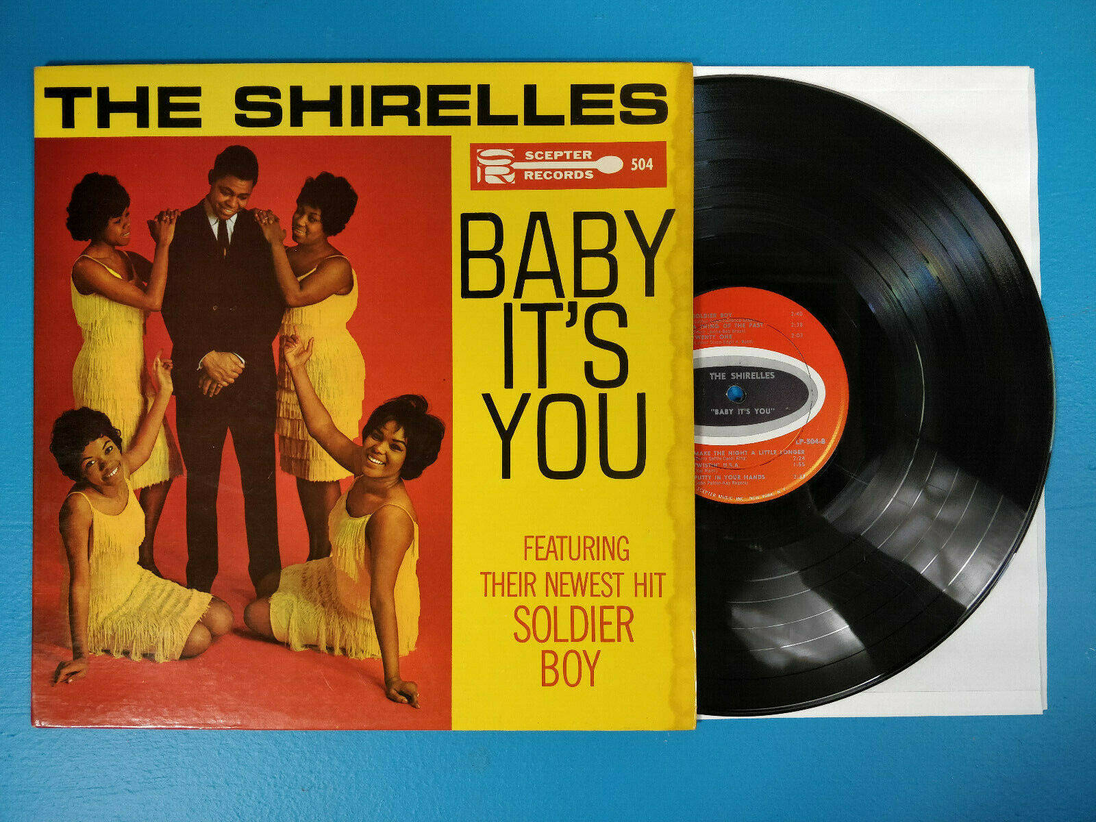vinyl 桌布 壁紙 — The Shirelles 的《Baby It's You》復古 1960 年代黑膠唱片，鮮黃色封面和黑色 LP 在藍色表面上。