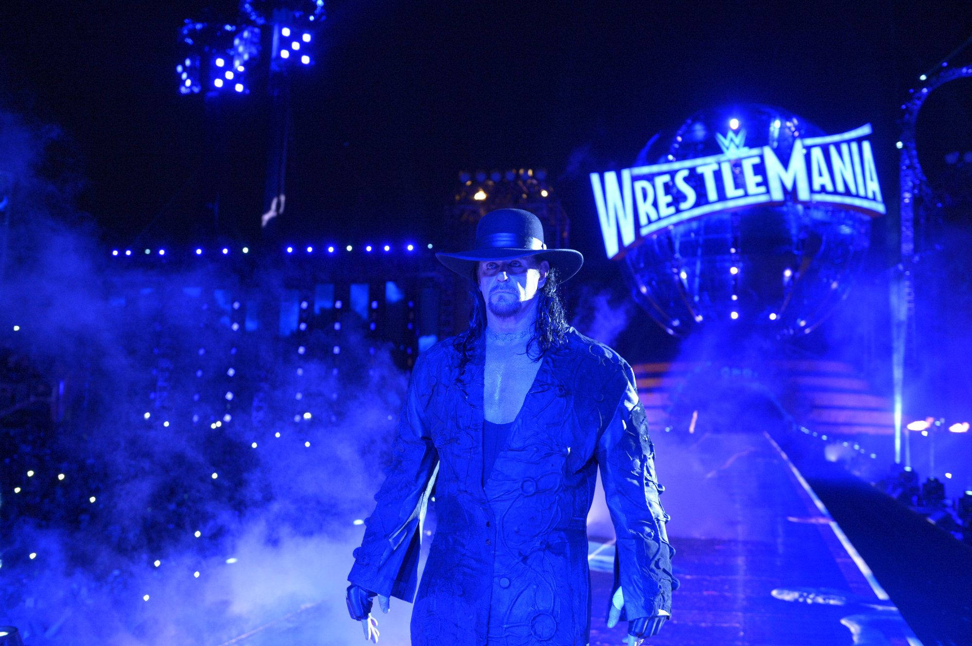 Undertaker 桌布 壁紙 — The Undertaker 在體育館燈光下，沿著藍色燈光的 WrestleMania 斜坡莊嚴地走下來，煙霧繚繞。