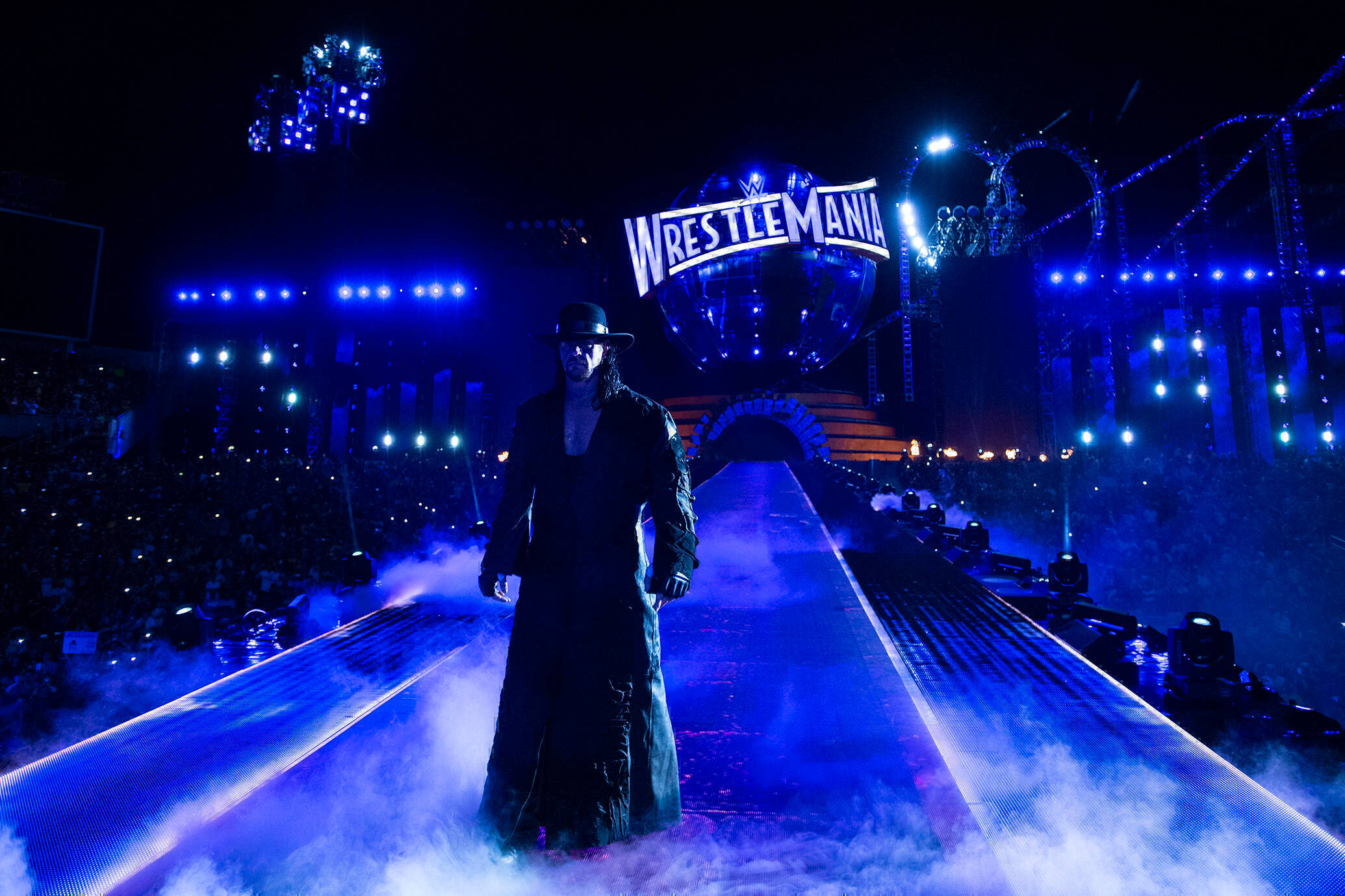 Undertaker 桌布 壁紙 — The Undertaker 沿著藍色燈光的 WrestleMania 斜坡走下來，周圍環繞著霧氣和體育場燈光。