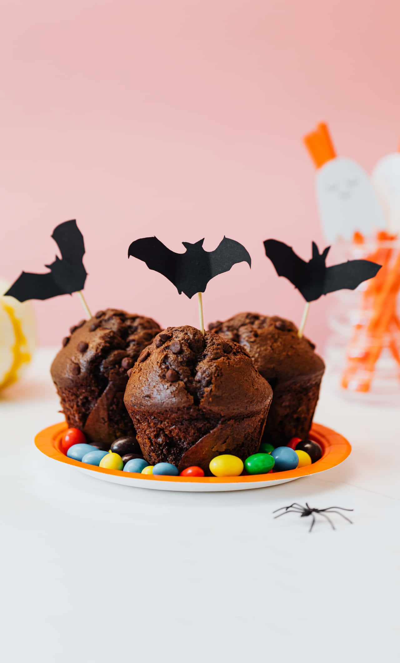 Halloween Treats 桌布 壁紙 — 三個巧克力碎片鬆餅，上面插有蝙蝠裝飾棒和彩色糖果，放在粉紅色背景的盤子上。