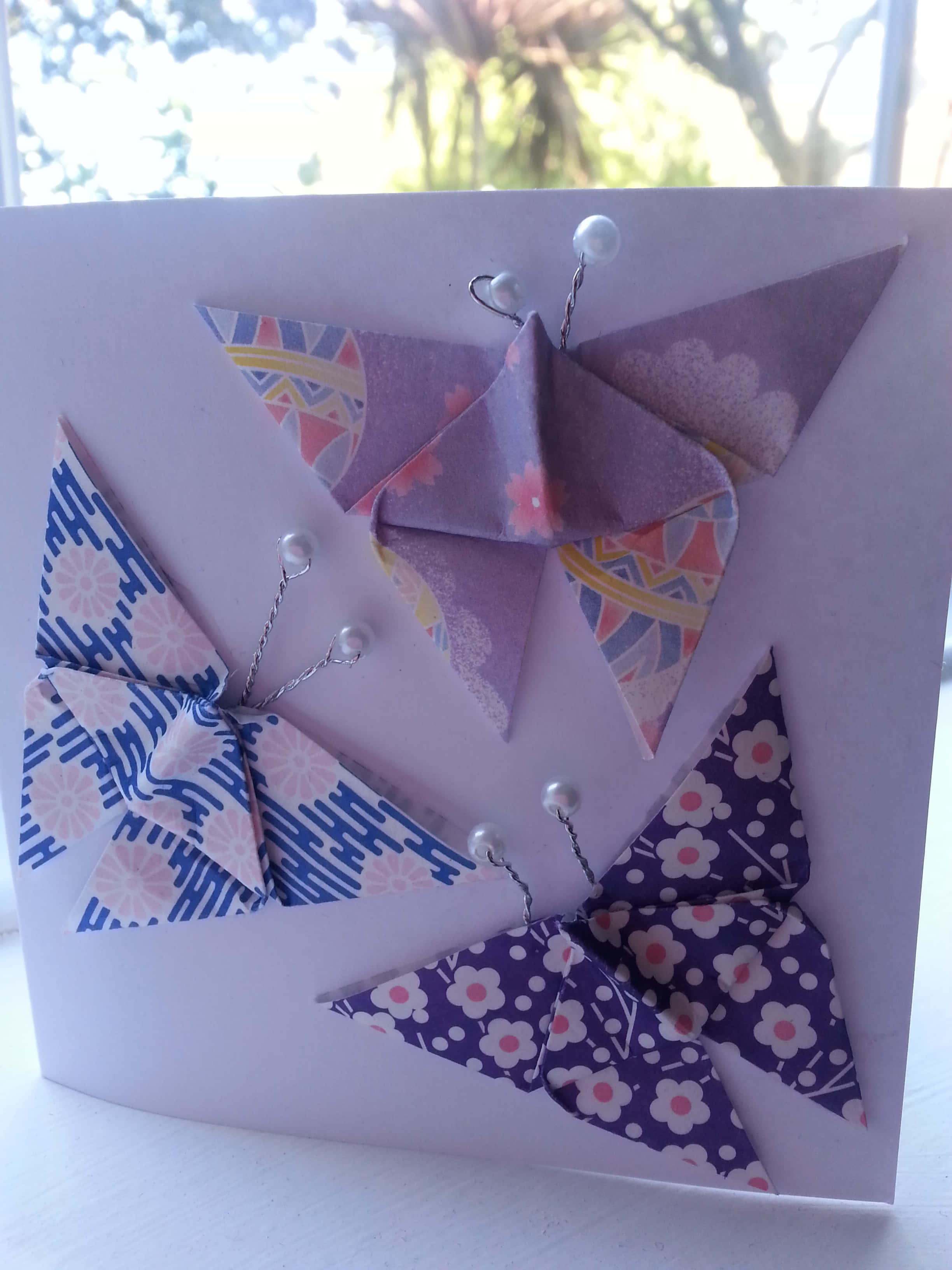 Butterfly Origami 桌布 壁紙 — 三隻色彩繽紛的摺紙蝴蝶用珍珠頭大頭針固定在薰衣草色卡片上，背景是明亮的窗戶。