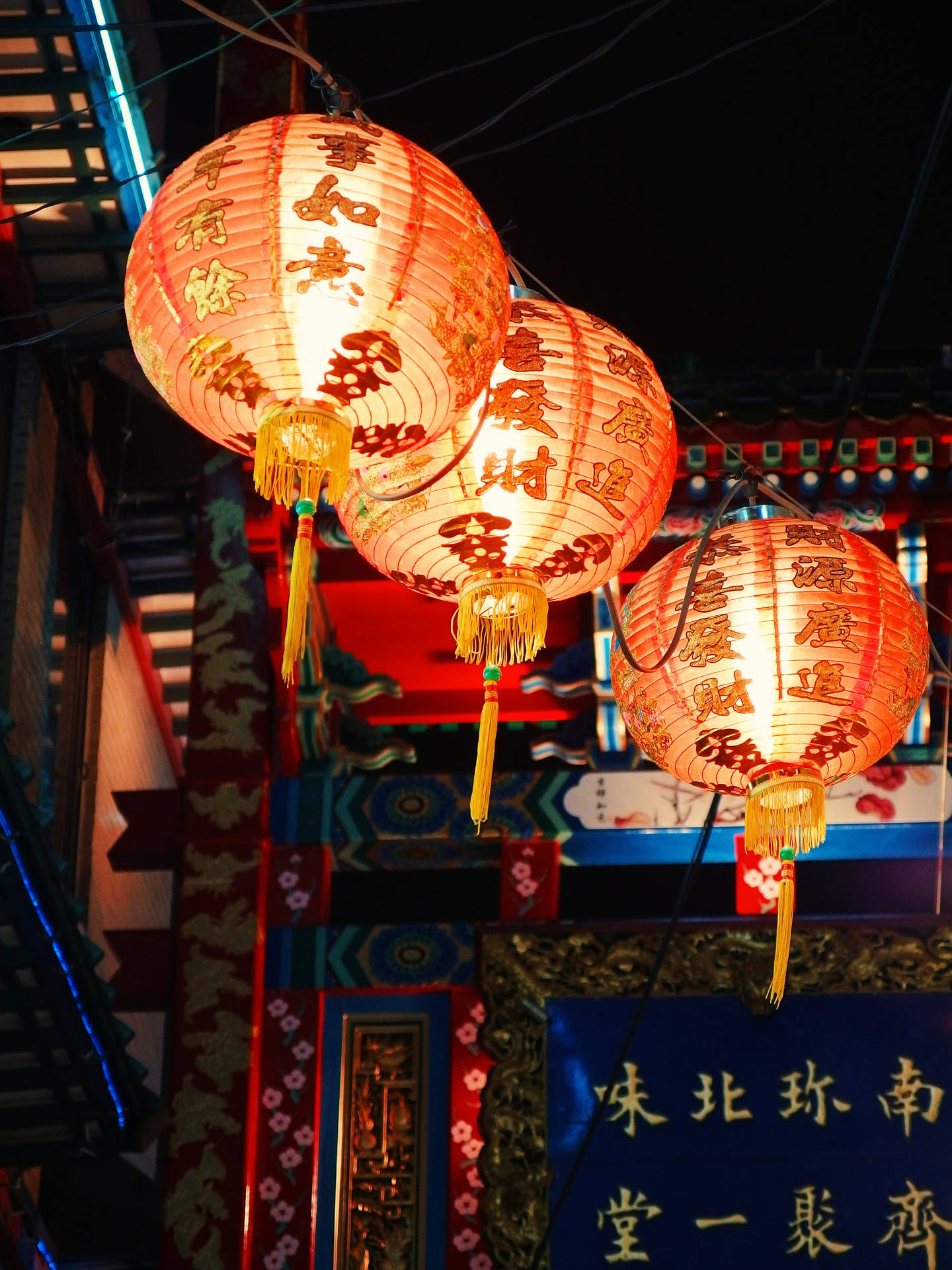 三個照亮的中國紅色燈籠，帶有金色流蘇，夜晚懸掛在傳統建築上方。 — 中國 壁紙
