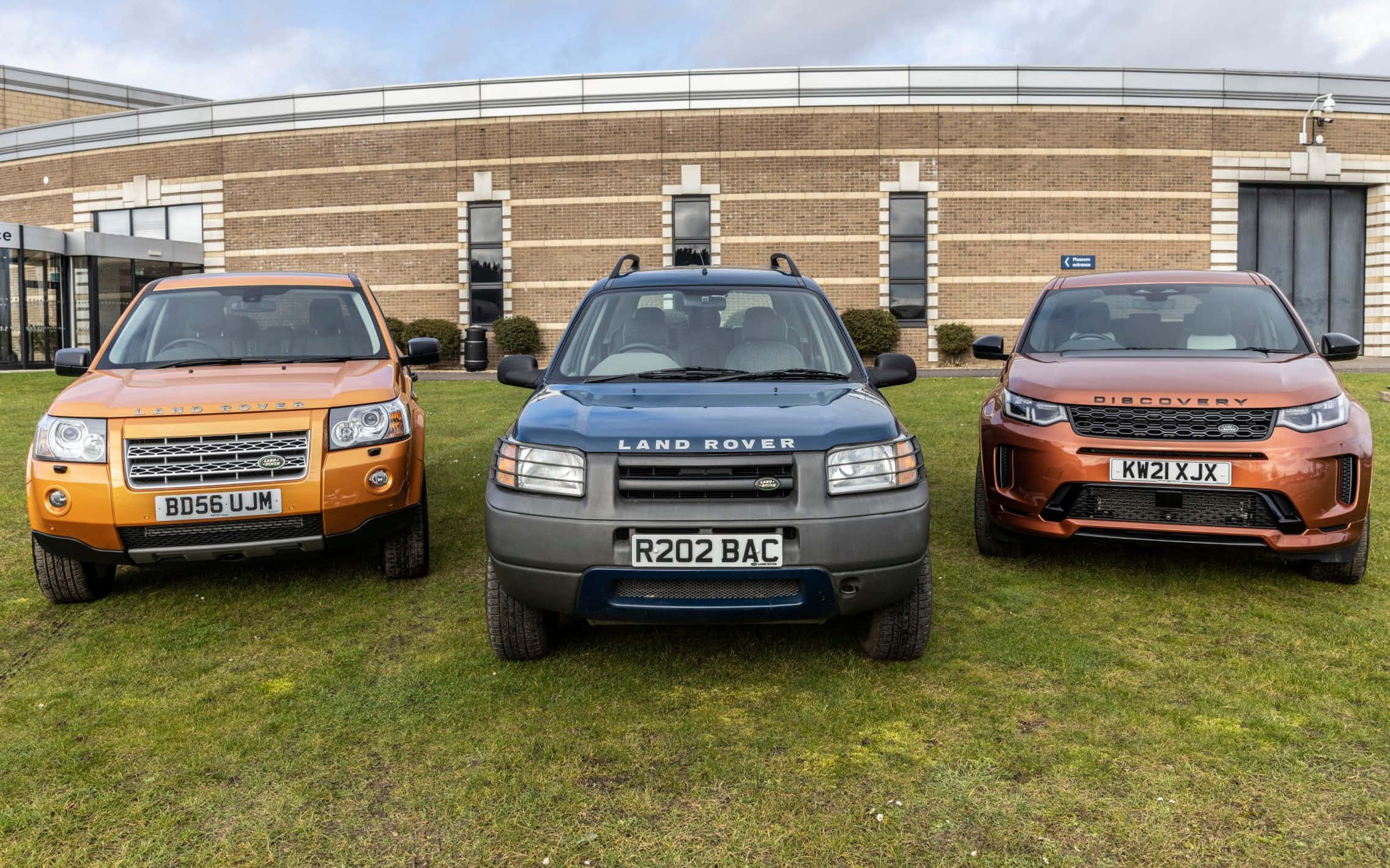 Land Rover Freelander 桌布 壁紙 — 三輛橙色、藍色和銅色的 Land Rover SUV 停放在磚造建築前的草地上，天空多雲。