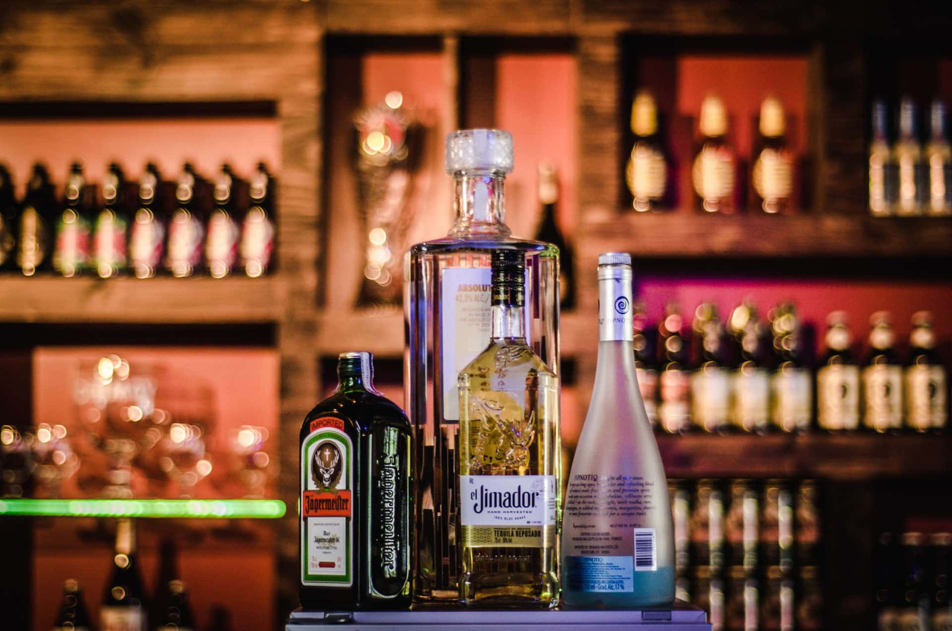  壁紙 — 三瓶酒類飲品醒目地擺放在吧檯上，背景是溫暖發光的酒瓶架。