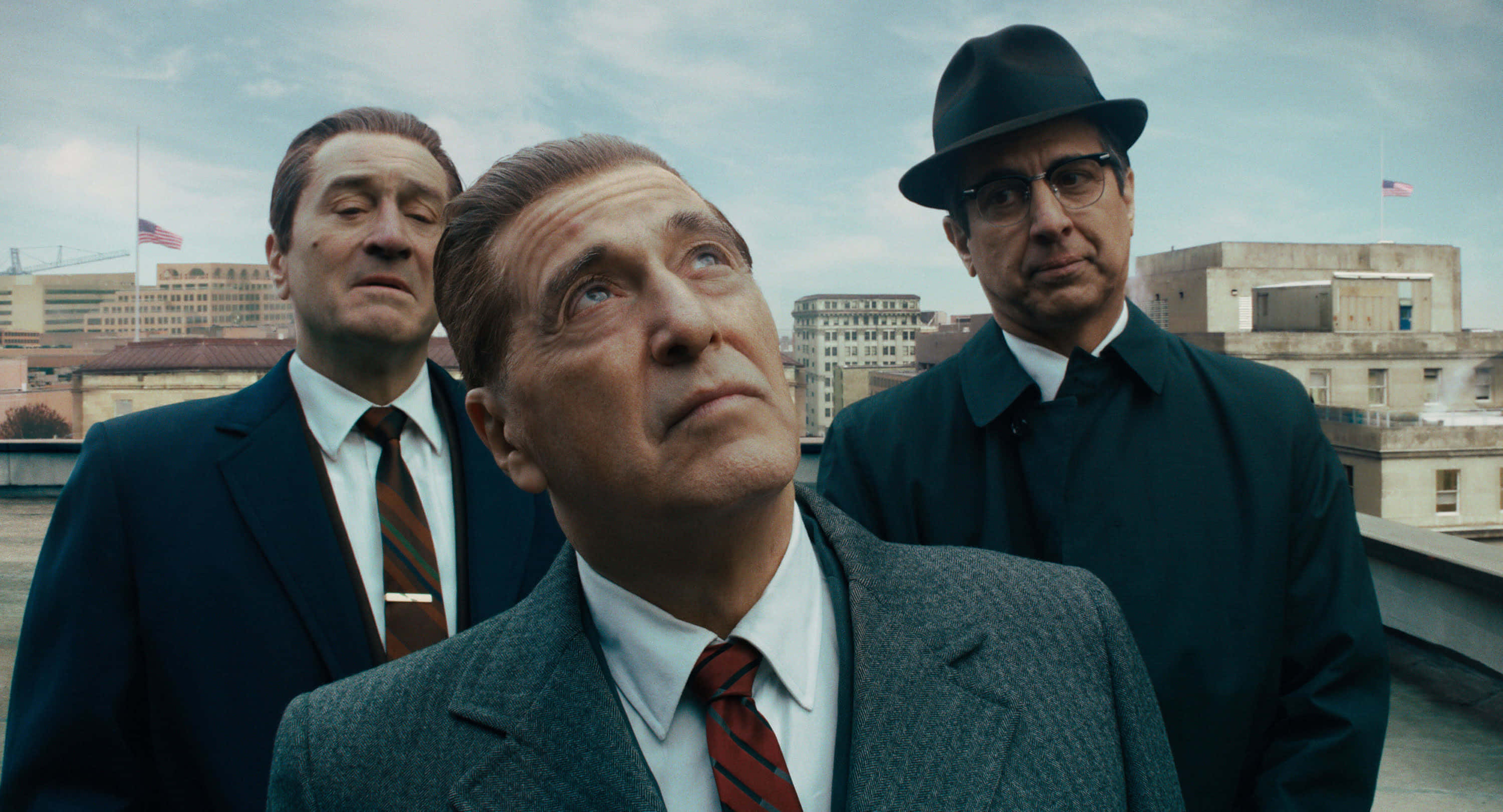 The Irishman 桌布 壁紙 — 三名身穿西裝戴帽子的男子站在屋頂上，表情嚴肅，背景是陰雲密佈的城市天際線。