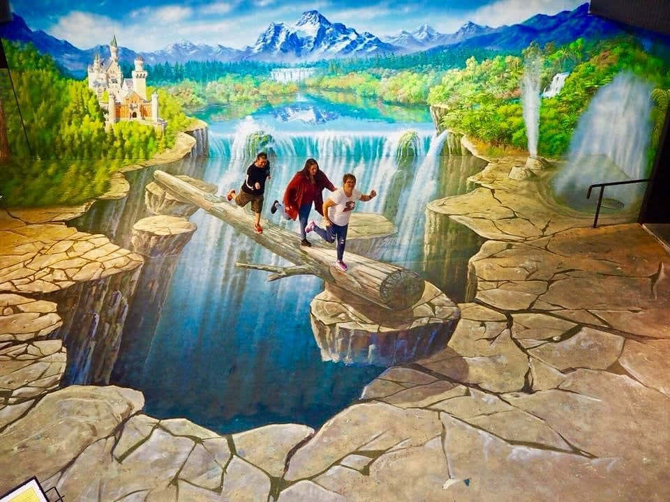 三個人跑過一條畫在峽谷上的木頭，旁邊有瀑布和山脈。 — 3D 壁紙