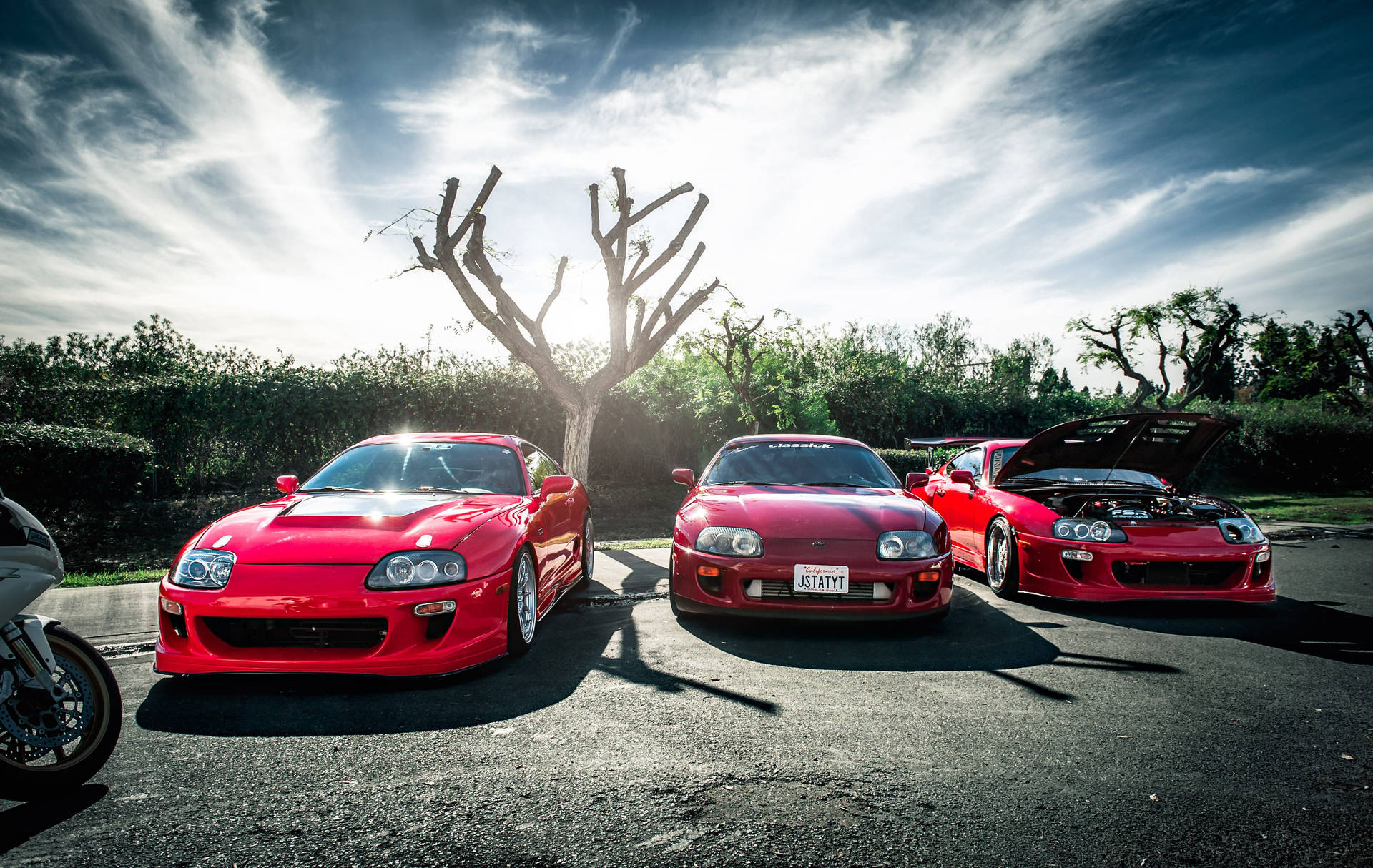 三輛紅色豐田Supra停放在柏油路上，有一棵光禿禿的樹和日落天空。 — Toyota Supra 壁紙