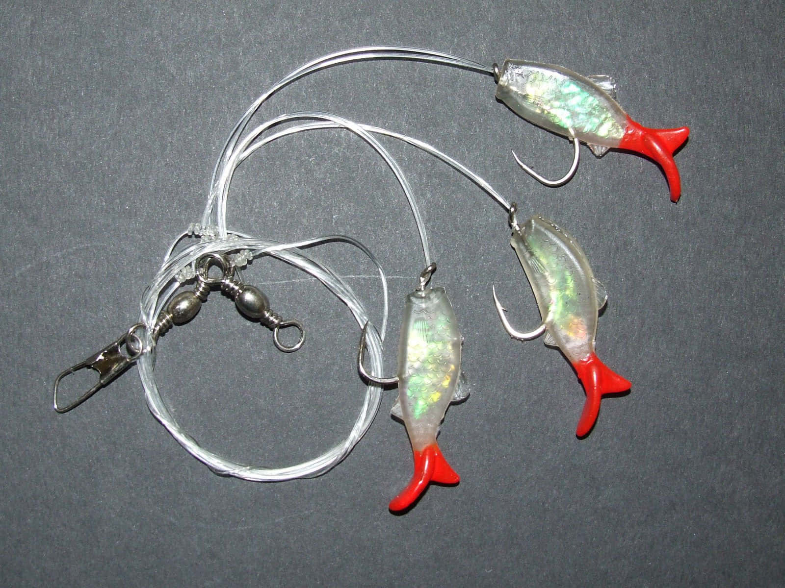  壁紙 — 三個閃亮銀色魚形假餌帶紅色尾巴，懸掛在透明魚線上，背景為深灰色。