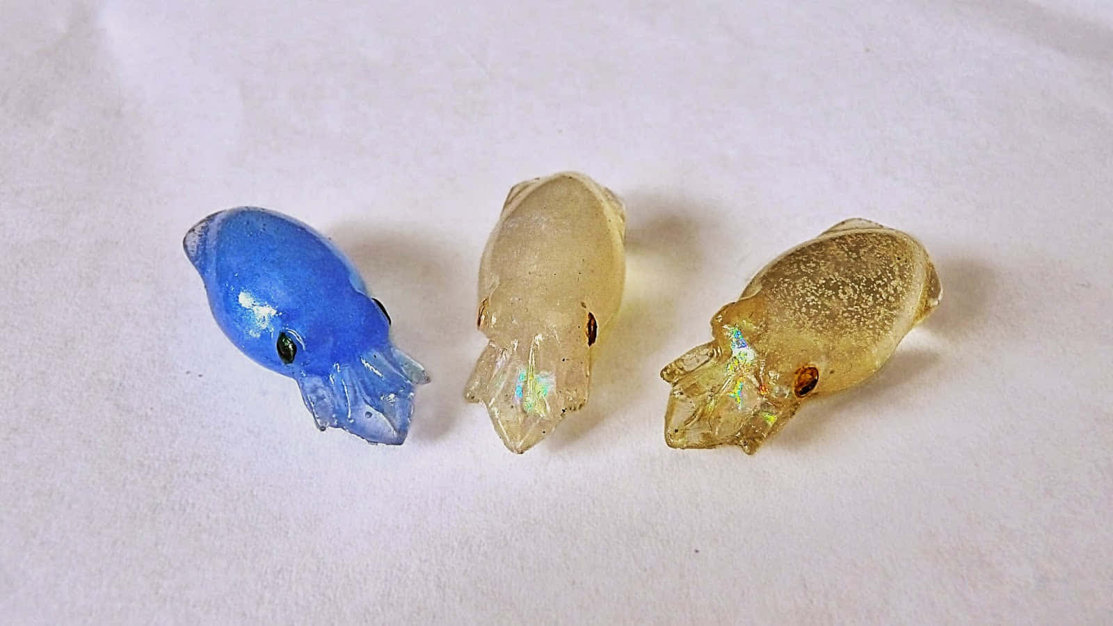  壁紙 — 三個藍色和琥珀色調的小玻璃魷魚雕像放在純白色表面上，捕捉光線，散發出彩虹般的光澤。