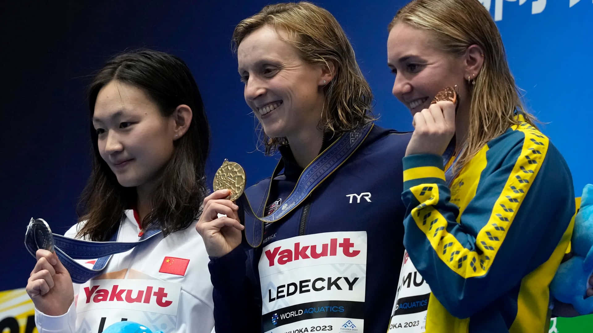  壁紙 — 三位微笑的女性游泳選手在世界游泳錦標賽活動中站在頒獎台上手持獎牌。