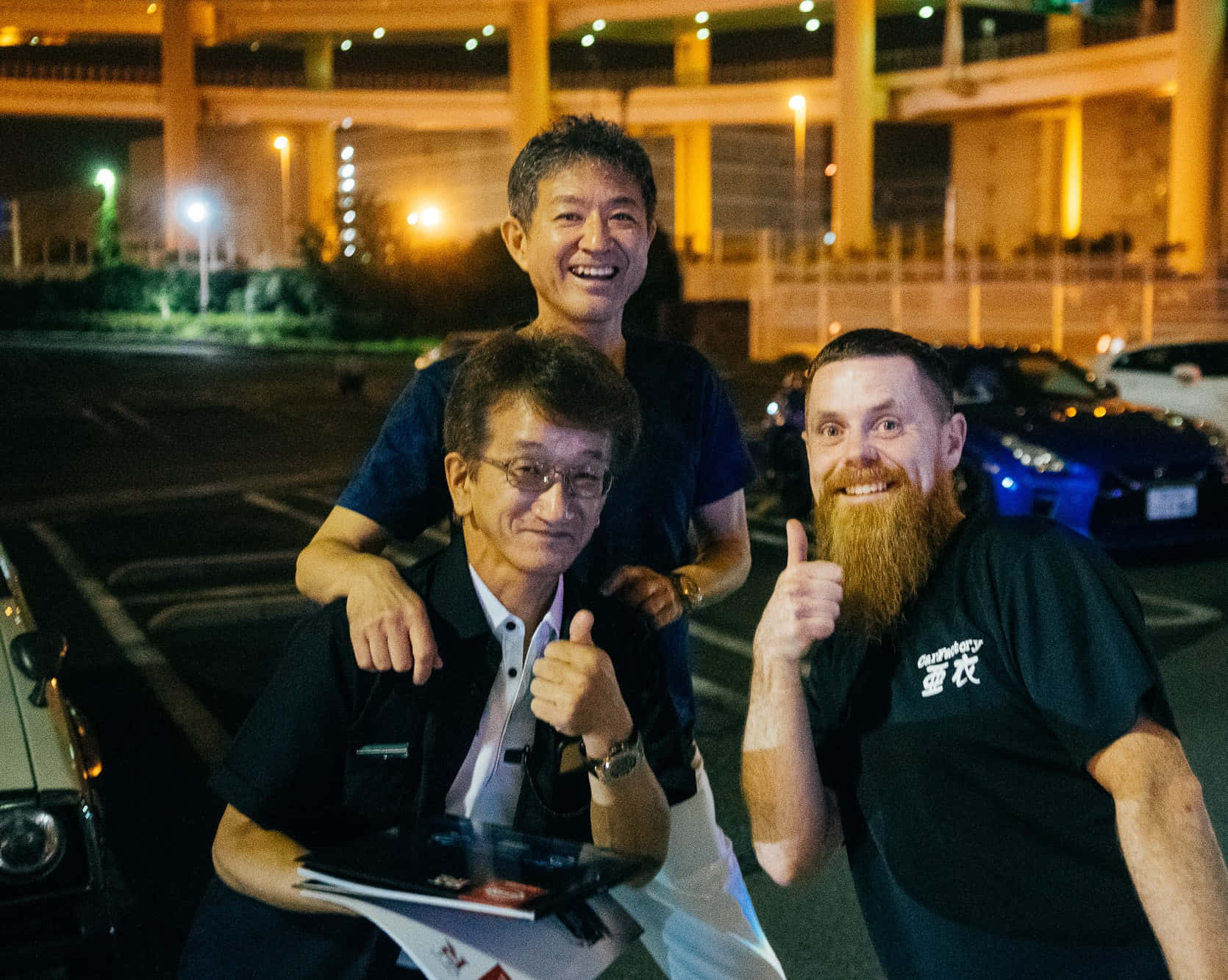  壁紙 — 三個微笑的男人在夜間停車場，在發光的街燈下，背後有汽車和天橋，愉快地擺姿勢。