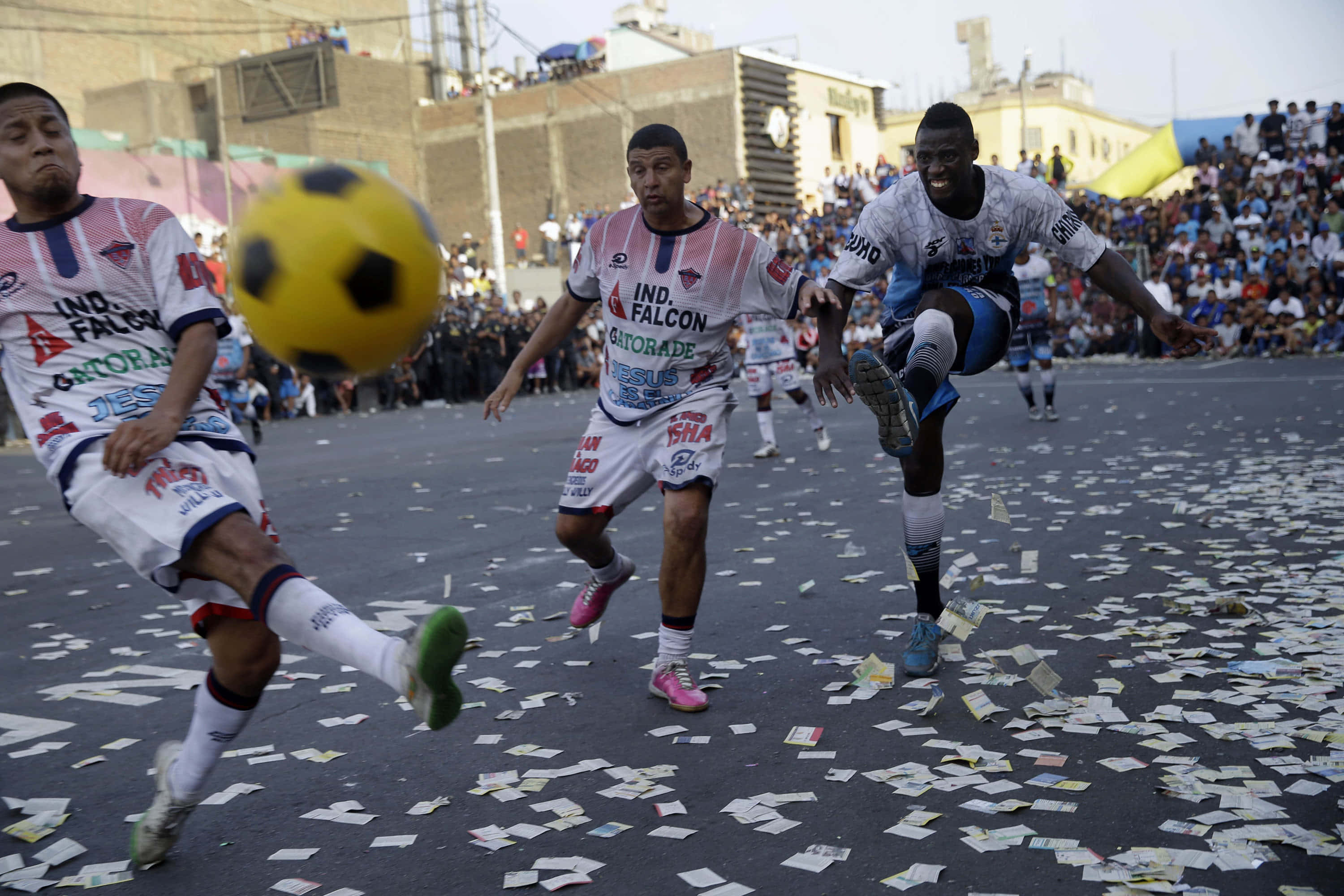 Street Soccer 桌布 壁紙 — 三名足球運動員在陽光普照、散落著五彩紙屑的街道上爭奪一個黃色足球。