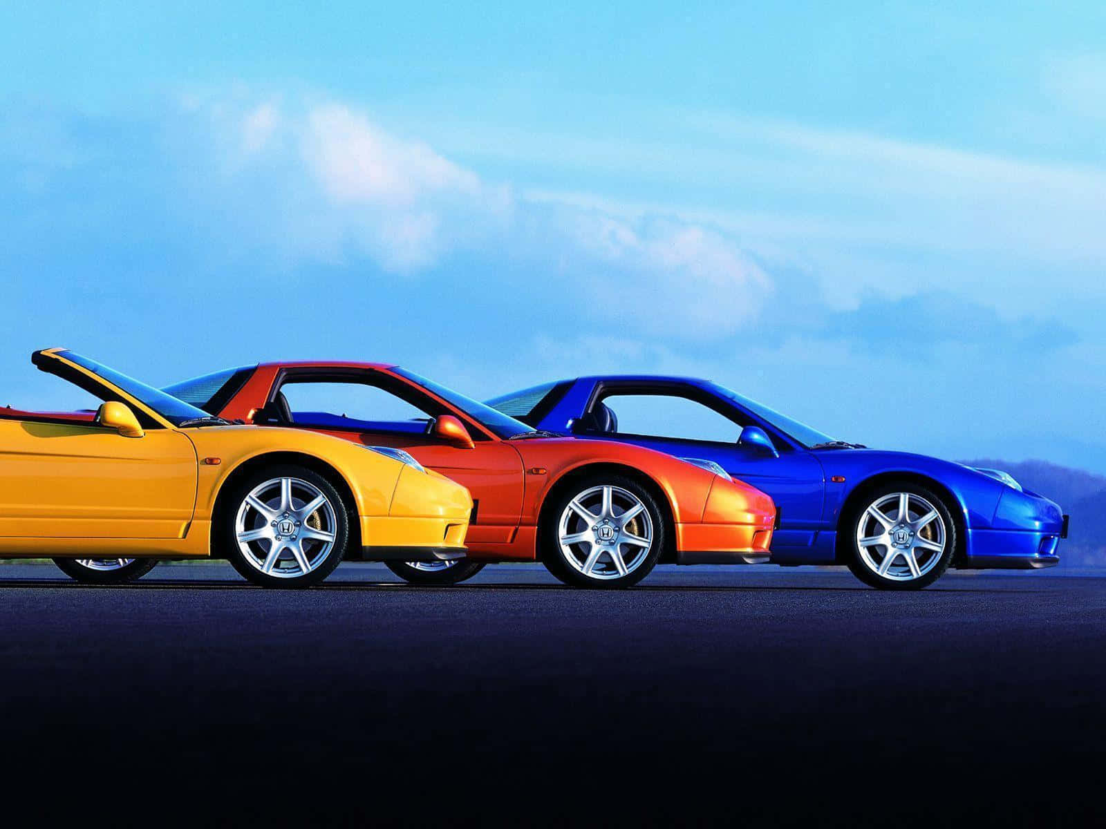 Acura Nsx 桌布 壁紙 — 三輛充滿活力的跑車，黃色、紅色和藍色，在柔和的藍天和縷縷白雲下，排列在一條黑暗的道路上。