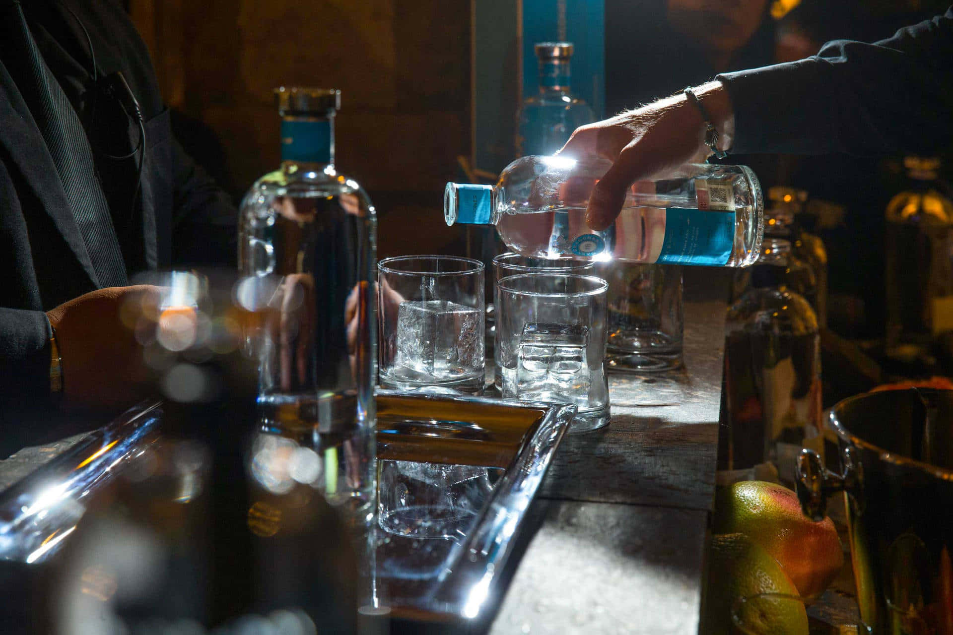  壁紙 — 調酒師在燈光昏暗的酒吧中，將發光的藍色利口酒倒入玻璃杯，附近有冰塊和水果。