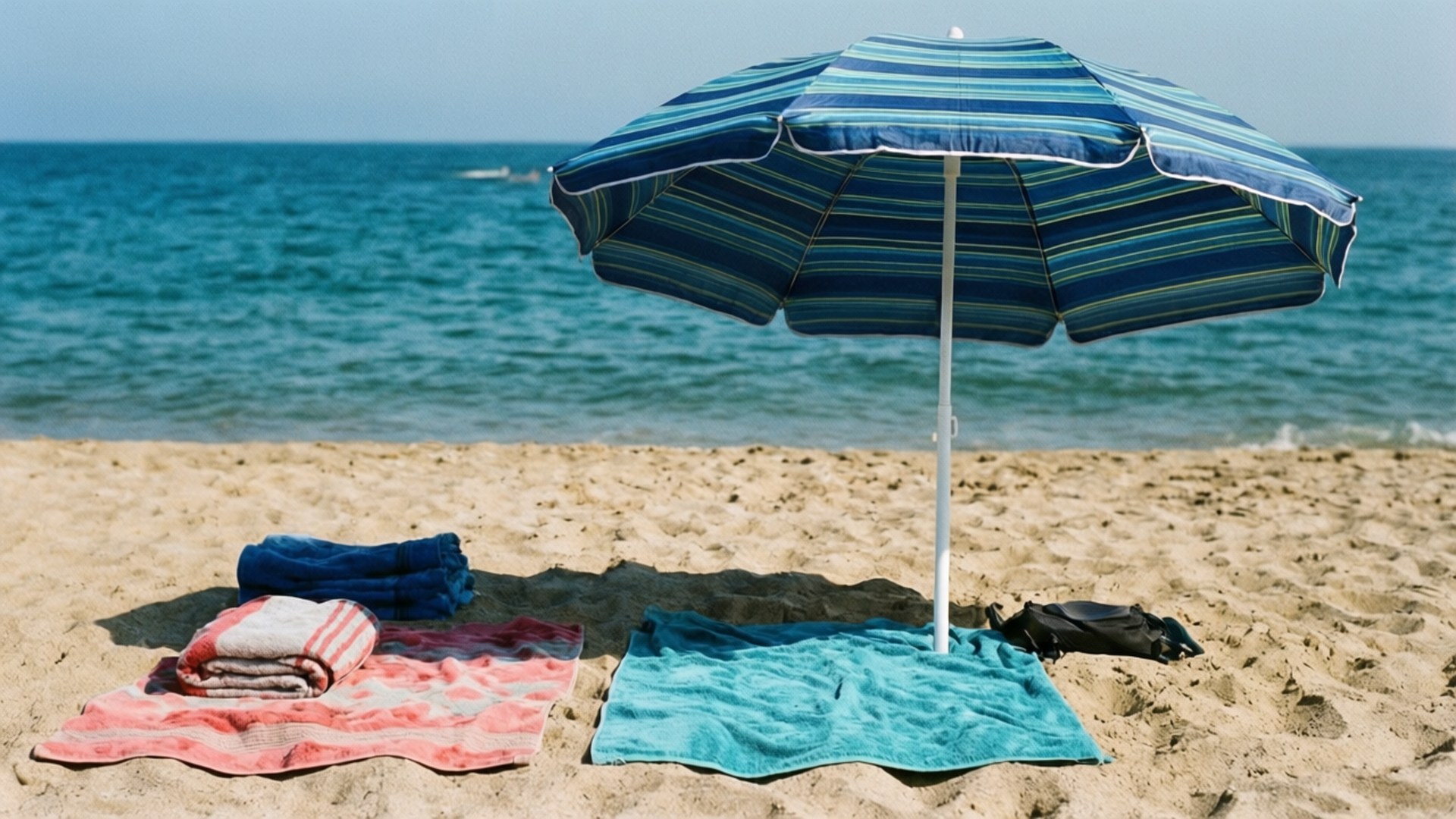 Beach Umbrella Haven 壁紙 — 一把條紋藍色沙灘傘在金色沙灘上為折疊的毛巾投下陰影，旁邊是碧綠的海浪。