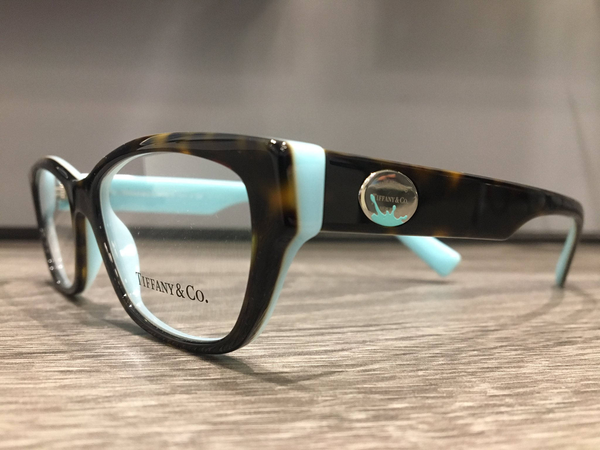 品牌 桌布 壁紙 — Tiffany & Co. 玳瑁眼鏡帶有綠松石點綴，置於木質表面上，柔和的工作室燈光下。