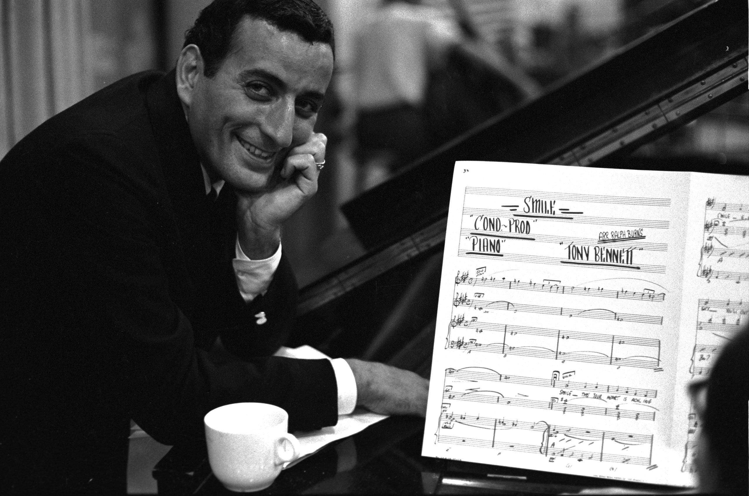 song 桌布 壁紙 — Tony Bennett 溫暖地微笑，倚靠在一架鋼琴上，面前放著打開的《Smile》樂譜。