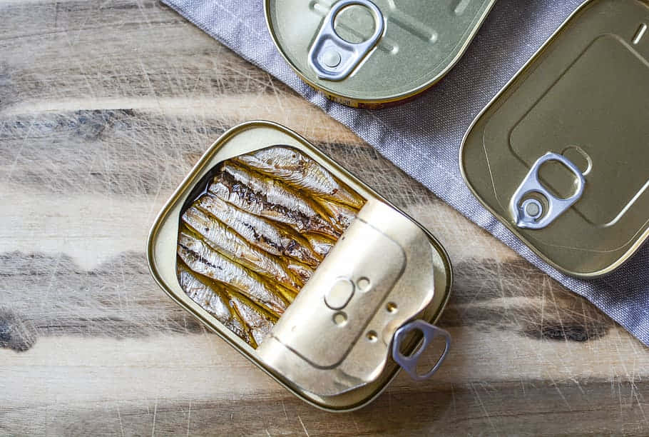  壁紙 — 俯視角度下，一個打開的沙丁魚罐頭放在木質表面上，附近有兩個未開封的罐頭。