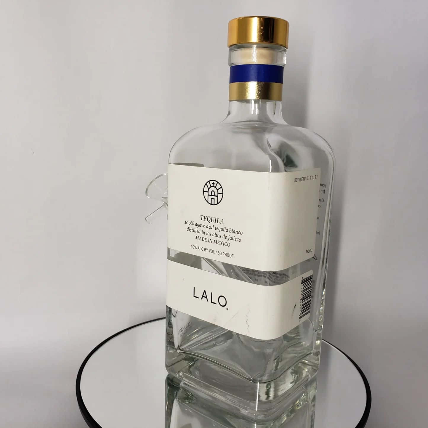  壁紙 — 一個透明玻璃龍舌蘭酒瓶，帶金色瓶蓋和白色標籤，放在鏡面托盤上，背景為純白色。