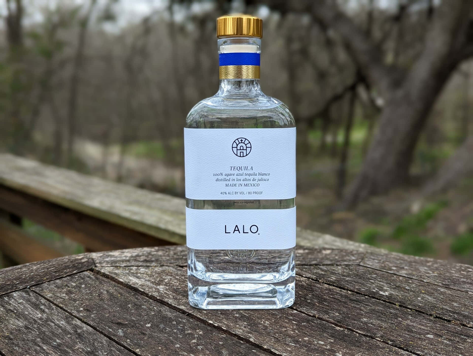  壁紙 — 一個透明的Lalo龍舌蘭酒瓶，金色瓶蓋，放在室外一張風化的木桌上，背景有模糊的樹木。