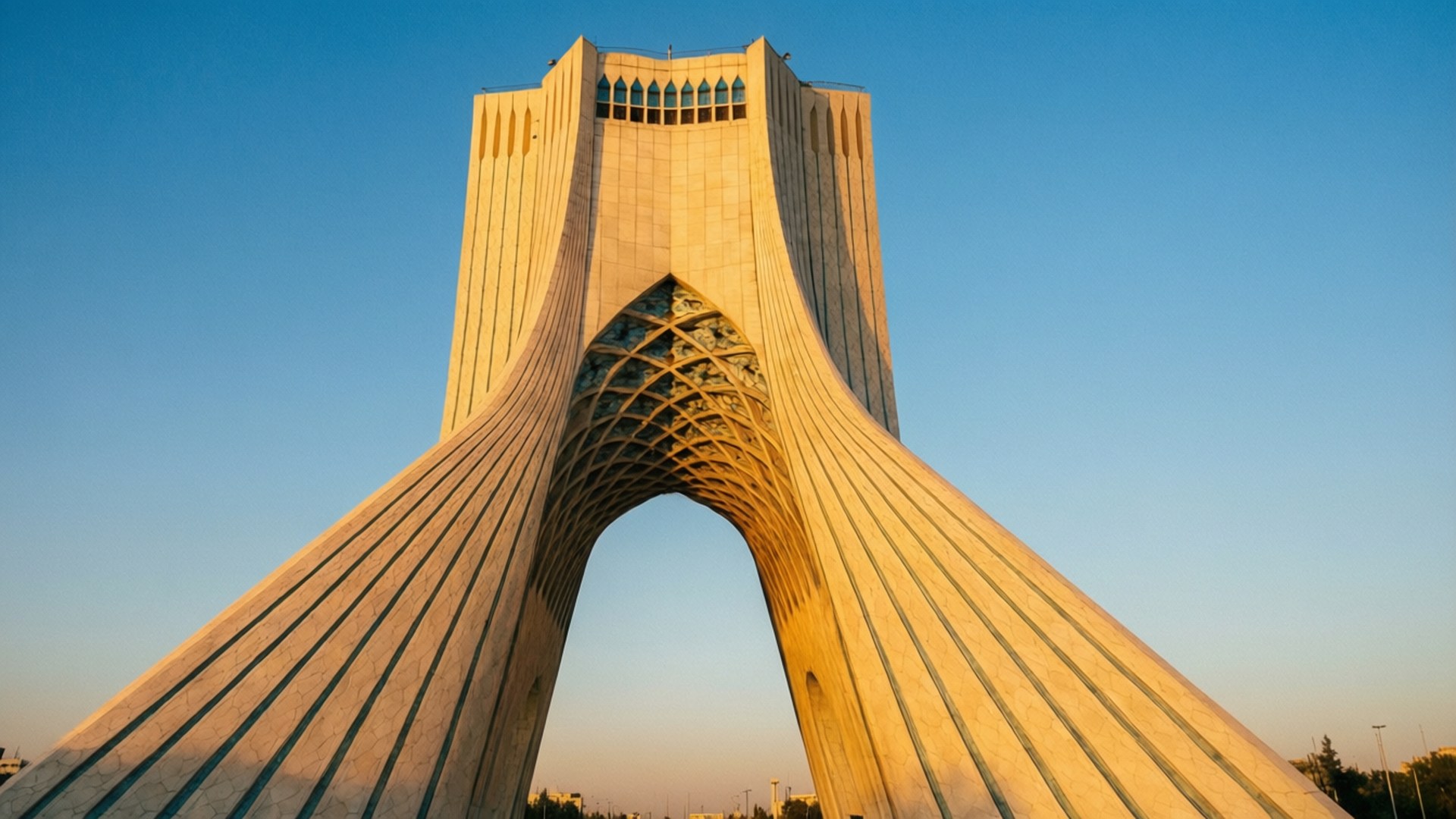 Azadi Tower Sunrise 壁紙 — 一座高聳的米色拱門紀念碑，帶有複雜的格子結構，在黃金時刻的清澈藍天下雄偉地矗立。
