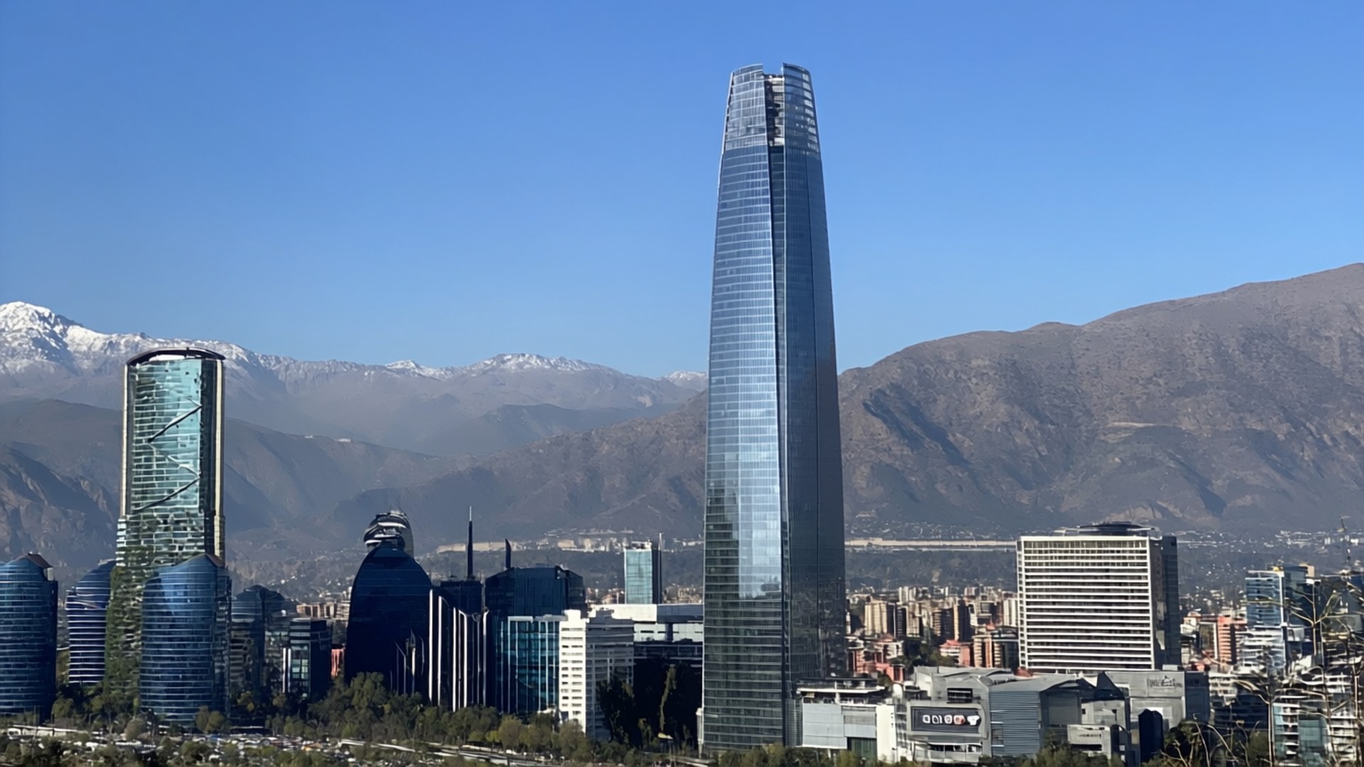 Santiago Cityscape 壁紙 — 高聳的玻璃摩天大樓刺穿清澈的藍天，背景中隱約可見白雪覆蓋的山峰。