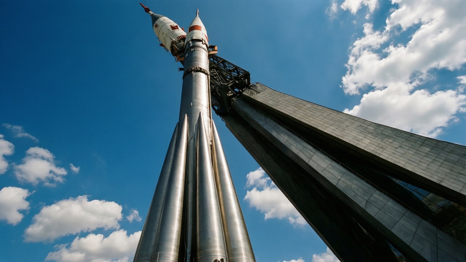 Space Monument 壁紙 — 一座高聳的銀色火箭紀念碑矗立在點綴著蓬鬆白雲的鮮豔藍天下。