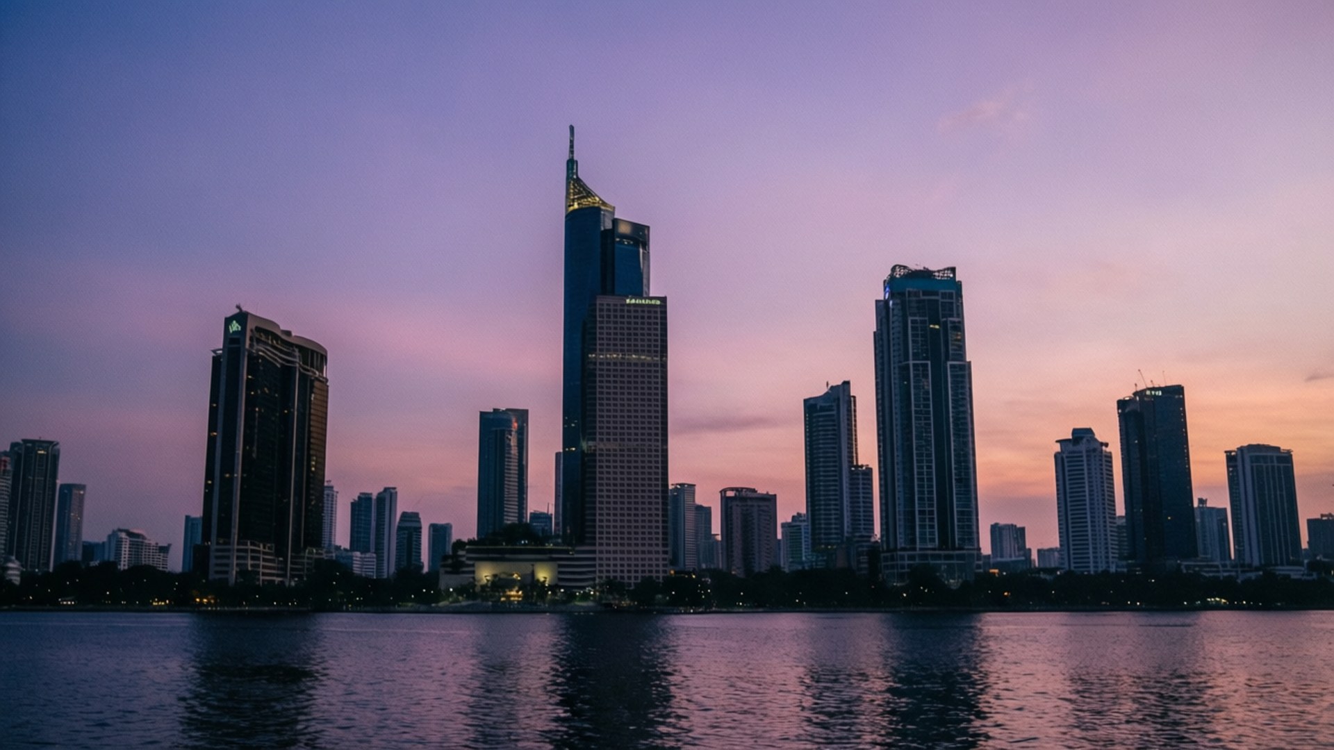 Dusk Skyline Silhouette 壁紙 — 高聳的摩天大樓在柔和的紫色和粉紅色暮色天空下倒映在平靜的水面上。