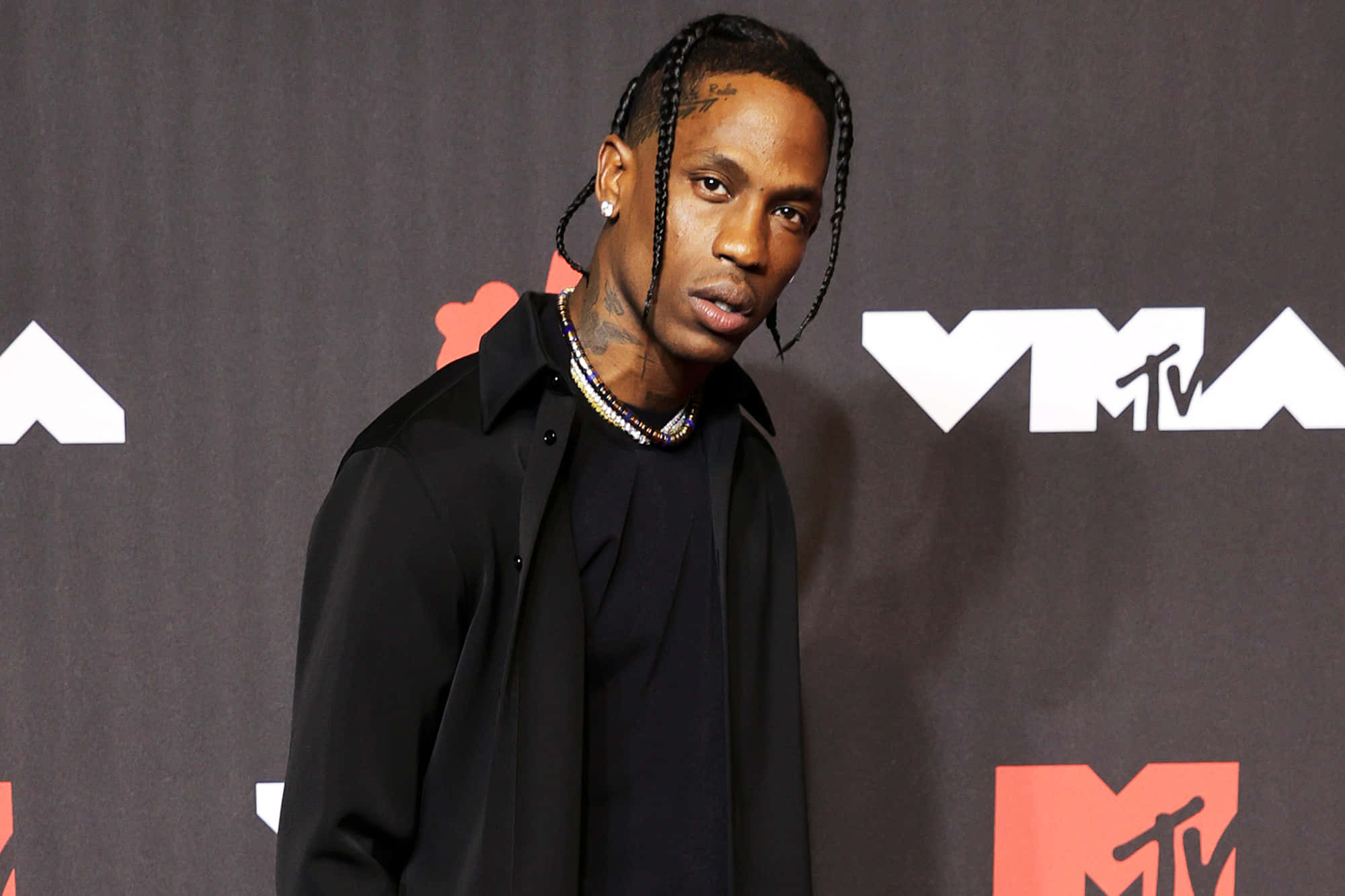 崔維斯·史考特 桌布 壁紙 — Travis Scott 自信地穿著黑色服裝，在帶有 MTV 標誌和橙色點綴的深色背景前擺姿勢。