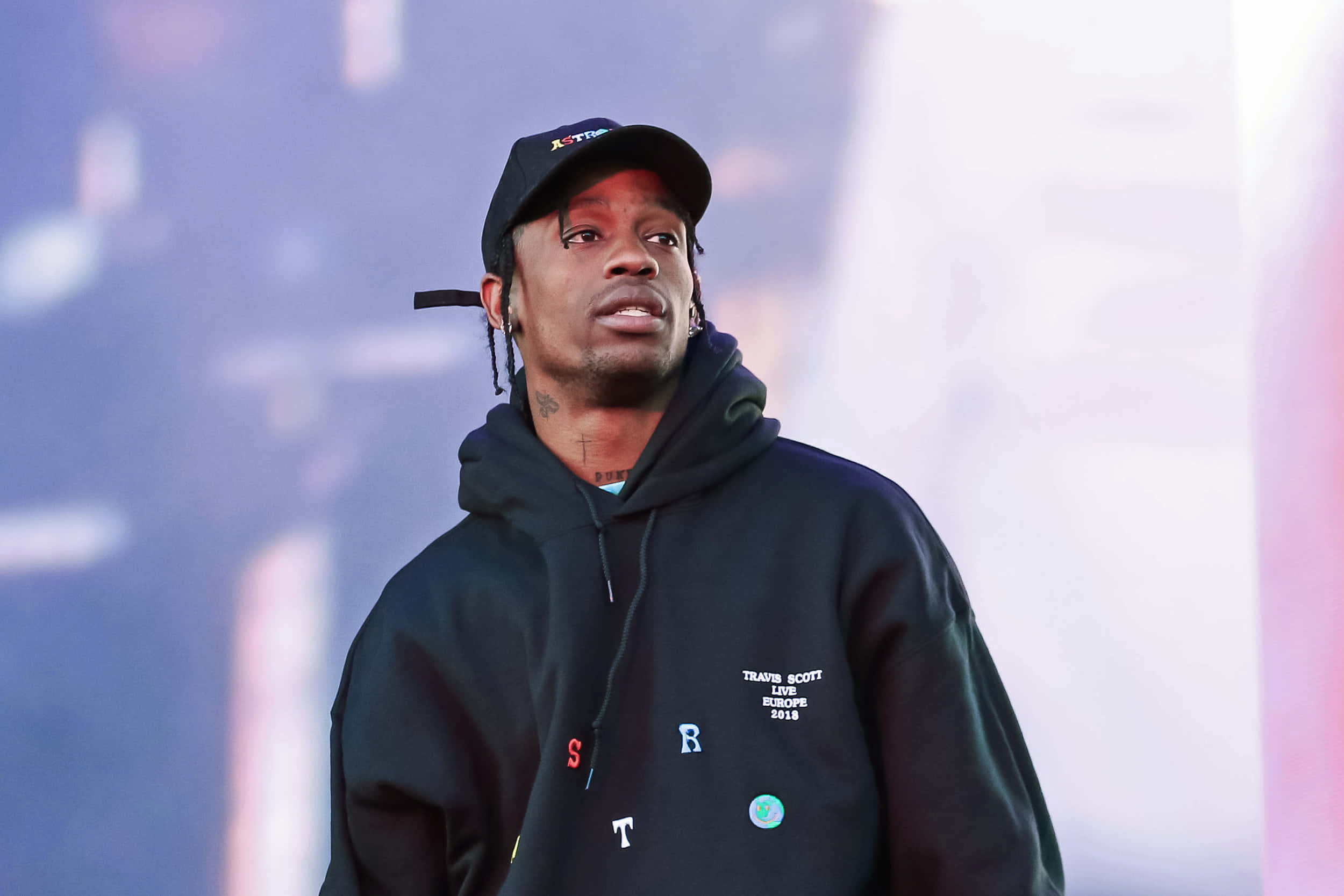 崔維斯·史考特 桌布 壁紙 — Travis Scott 身穿黑色連帽衫和帽子，在演唱會期間站在朦朧的紫色和粉紅色燈光下的舞台上。