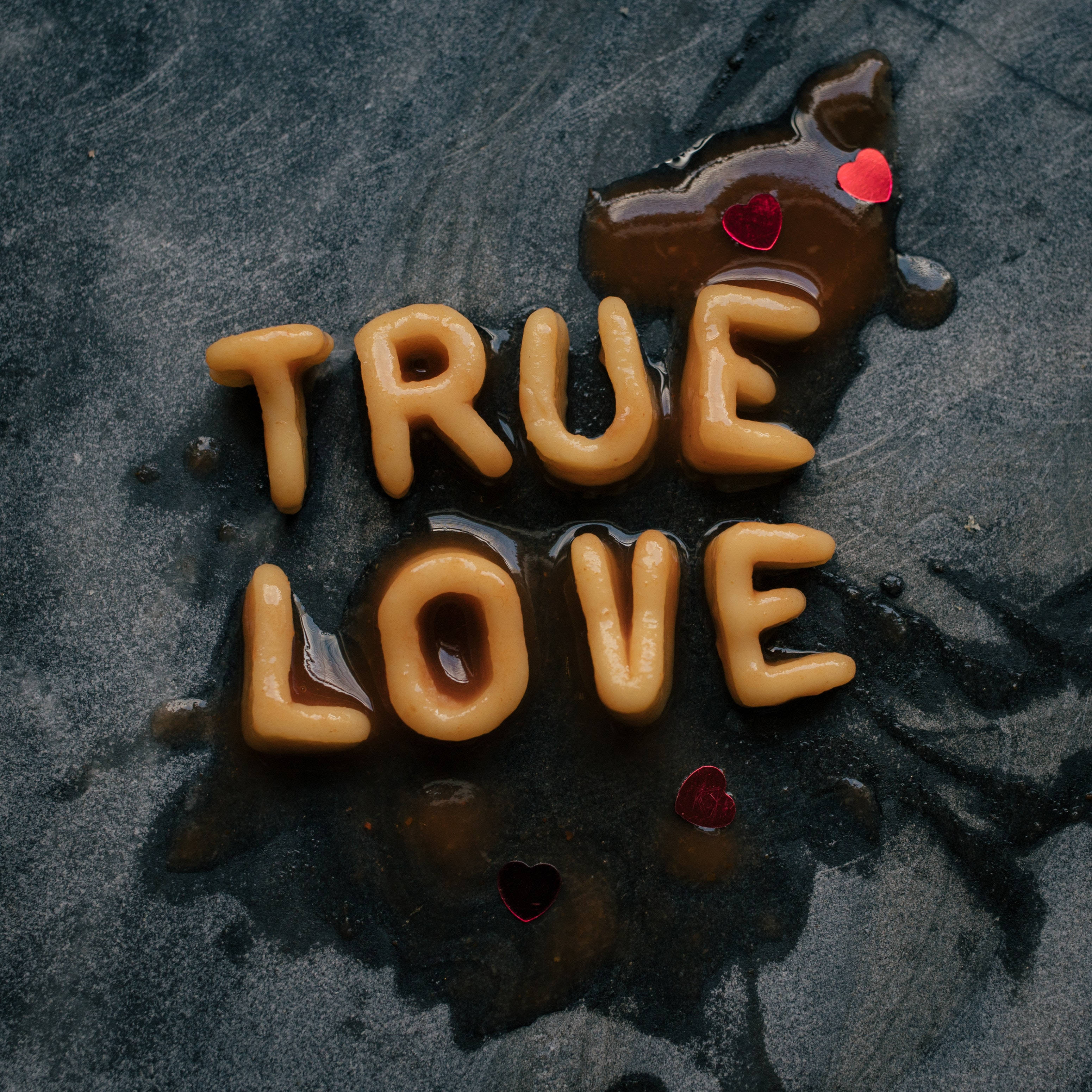 愛情語錄 桌布 壁紙 — 「true love」這幾個字用焦糖糖漿拼寫，上面有紅色心形糖果，在深色紋理表面上。