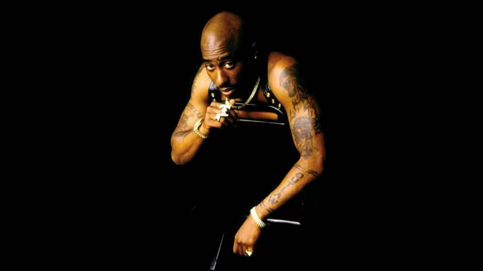 tupac 桌布 壁紙 — Tupac Shakur 在黑色背景前傾身，金色鏈條和紋身在戲劇性燈光下閃耀。