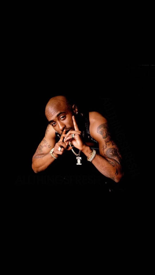 tupac 桌布 壁紙 — Tupac Shakur 在黑色背景前自信地擺姿勢，身上有刺青和珠寶，燈光效果戲劇化。