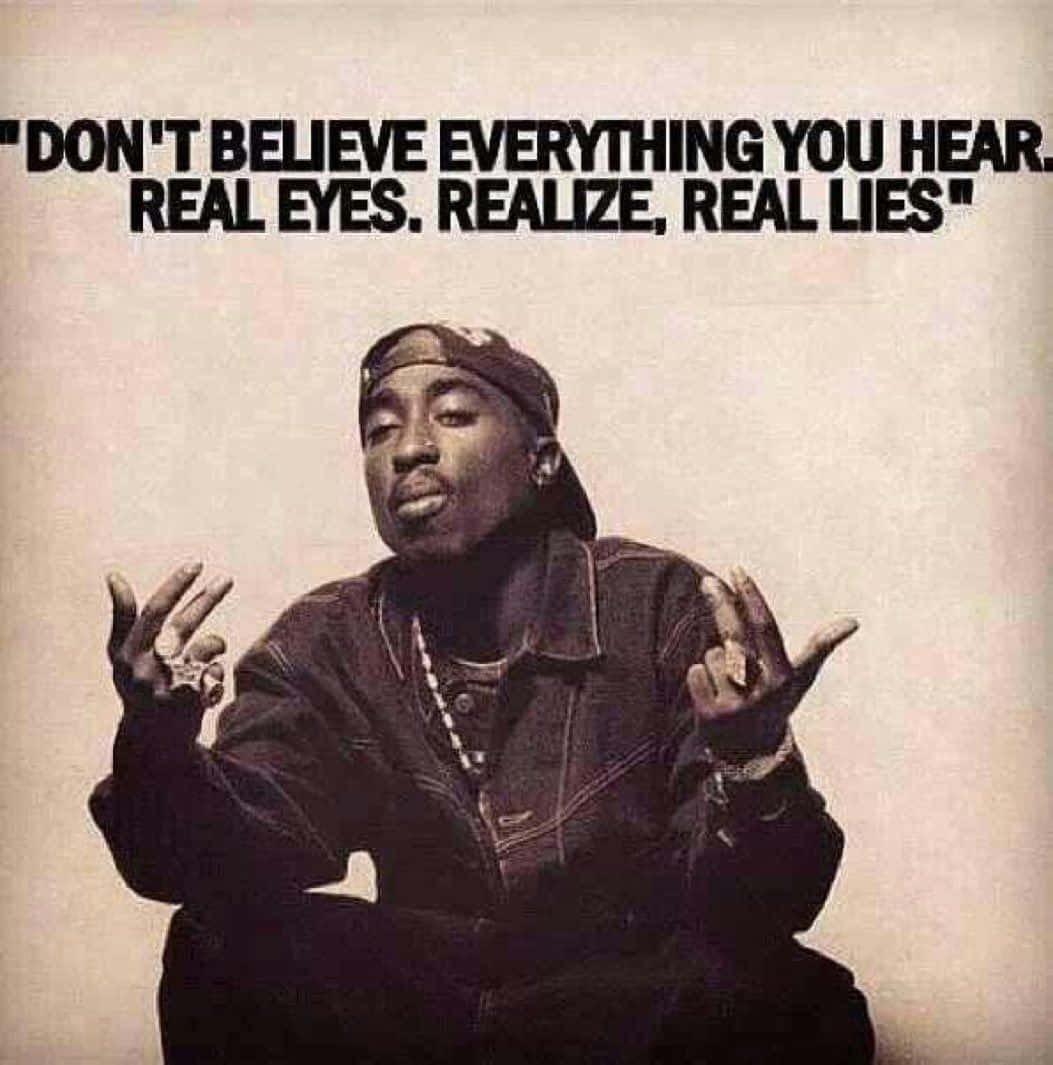 tupac 桌布 壁紙 — Tupac Shakur 坐在素色背景前，雙手舉起，戴著帽子和外套，上方有粗體文字寫著「DON'T BELIEVE EVERYTHING YOU HEAR REAL EYES REALIZE REAL LIES」。