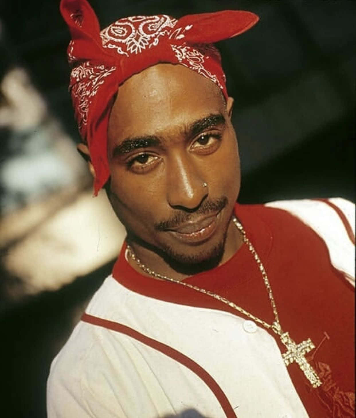 tupac 桌布 壁紙 — Tupac Shakur 戴著紅色頭巾和金色十字架項鍊，在黑暗模糊的背景前凝視著。