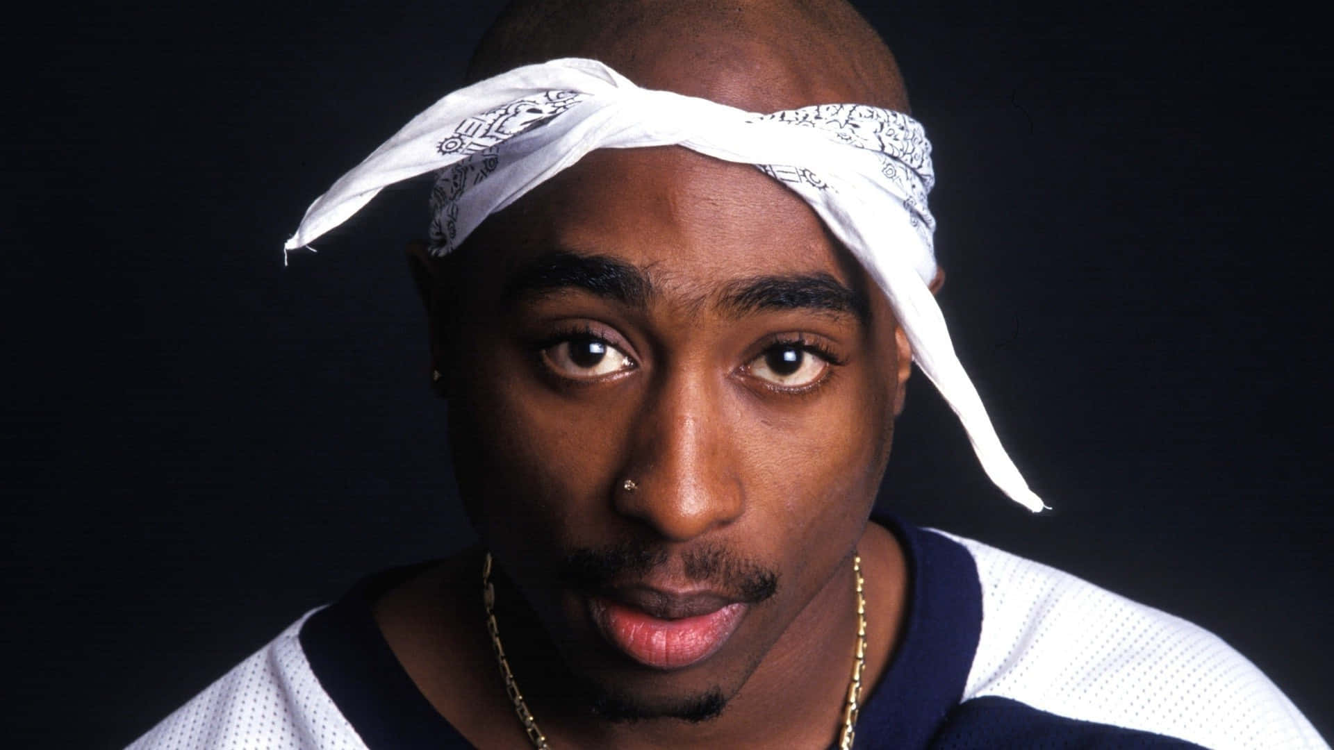 tupac 桌布 壁紙 — Tupac Shakur 的特寫肖像，戴著白色頭巾和金鏈，在黑色背景前強烈凝視。