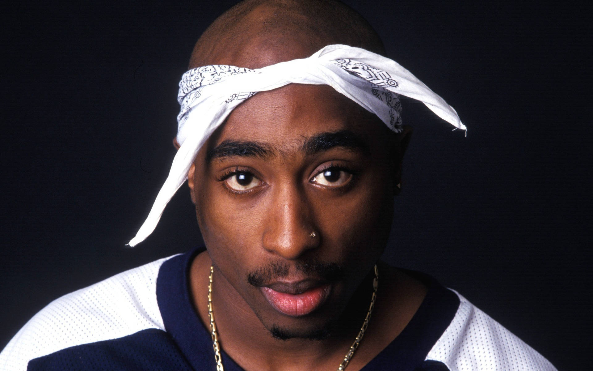 Tupac Shakur 戴著白色頭巾和金鏈，直視鏡頭。